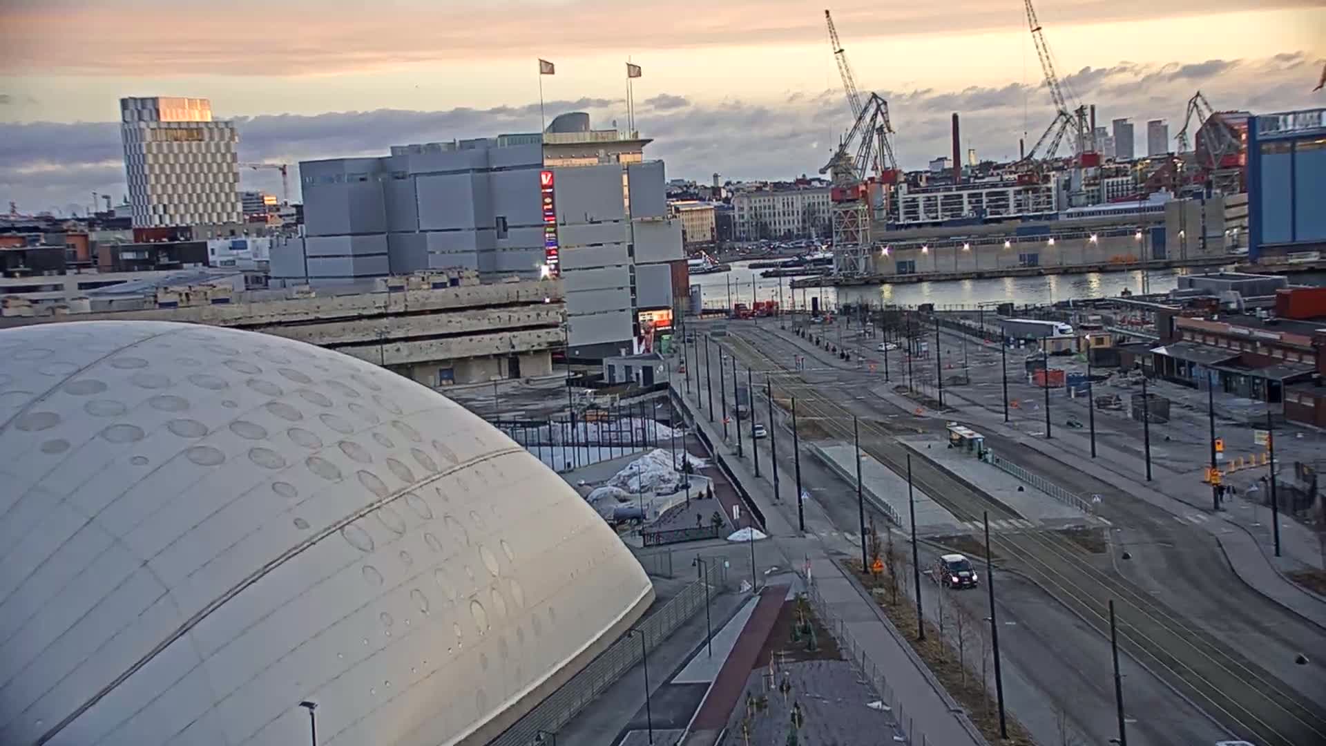 A large domed building sits beside a wide, mostly empty street with tram tracks, overlooking a harbor with several cranes under a partly cloudy sunset sky.