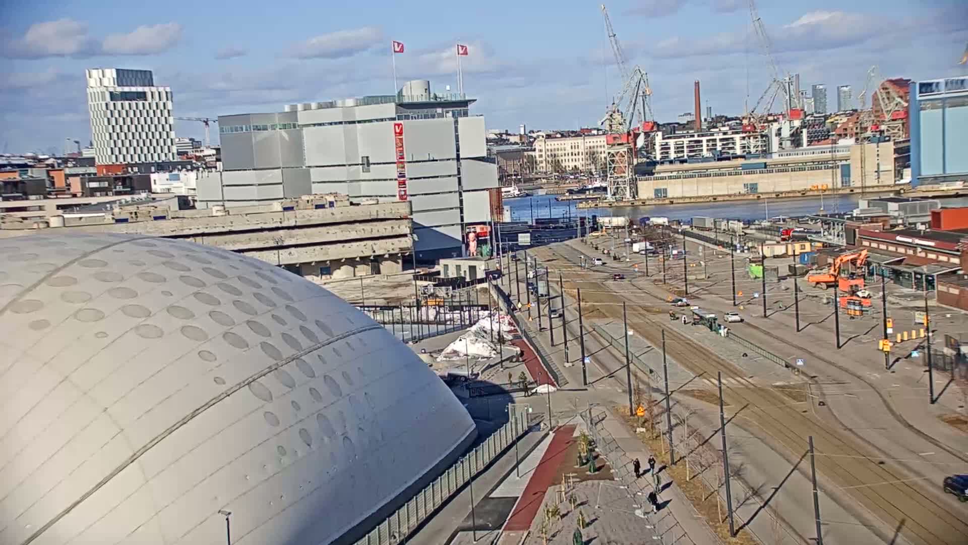 A large domed building is seen from above on a sunny day, with a wide street and harbor in the background, showing construction and various vehicles.