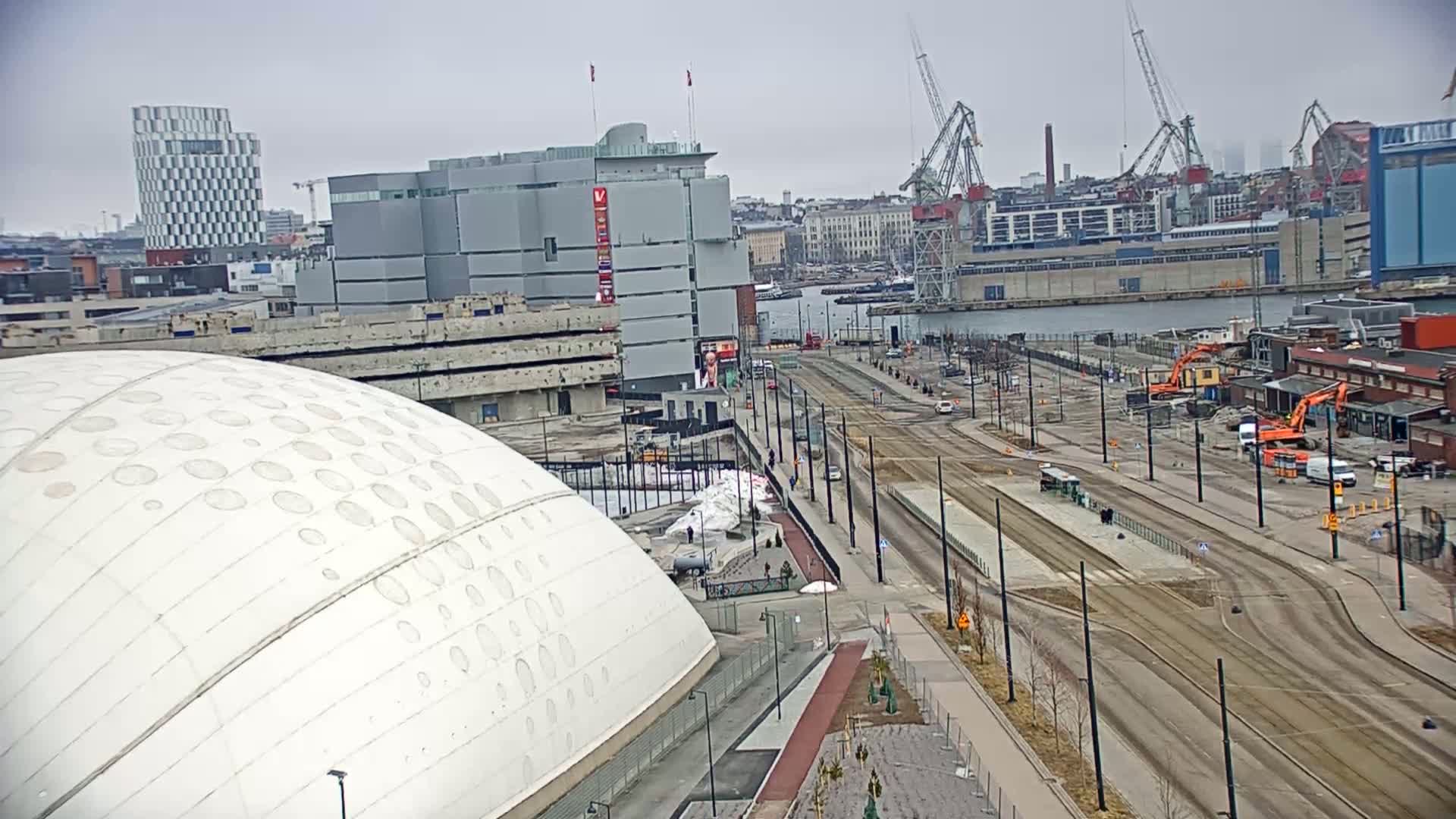 A large white dome-shaped building sits near a waterfront under an overcast sky, with construction and tram tracks visible in the foreground and cranes and industrial buildings in the background.