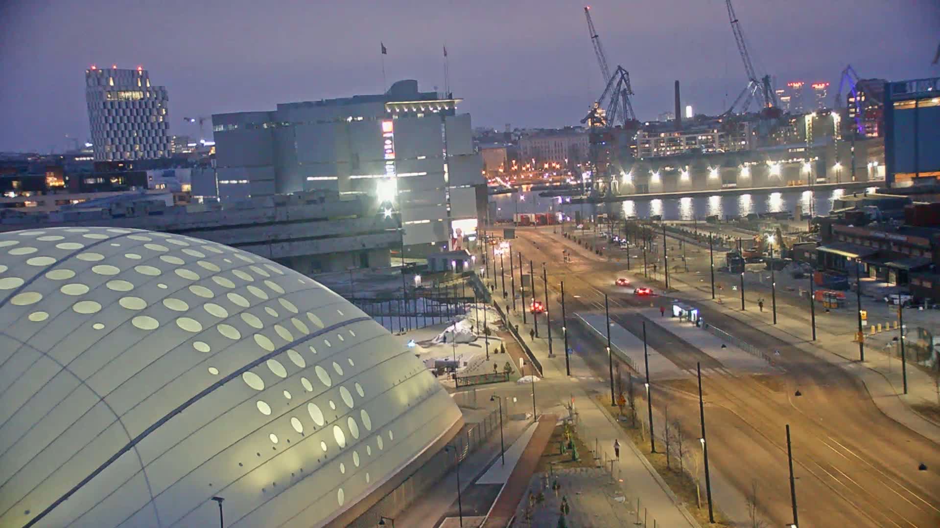 An illuminated domed building sits beside a city street at dusk, with a harbor and cranes visible in the background.
