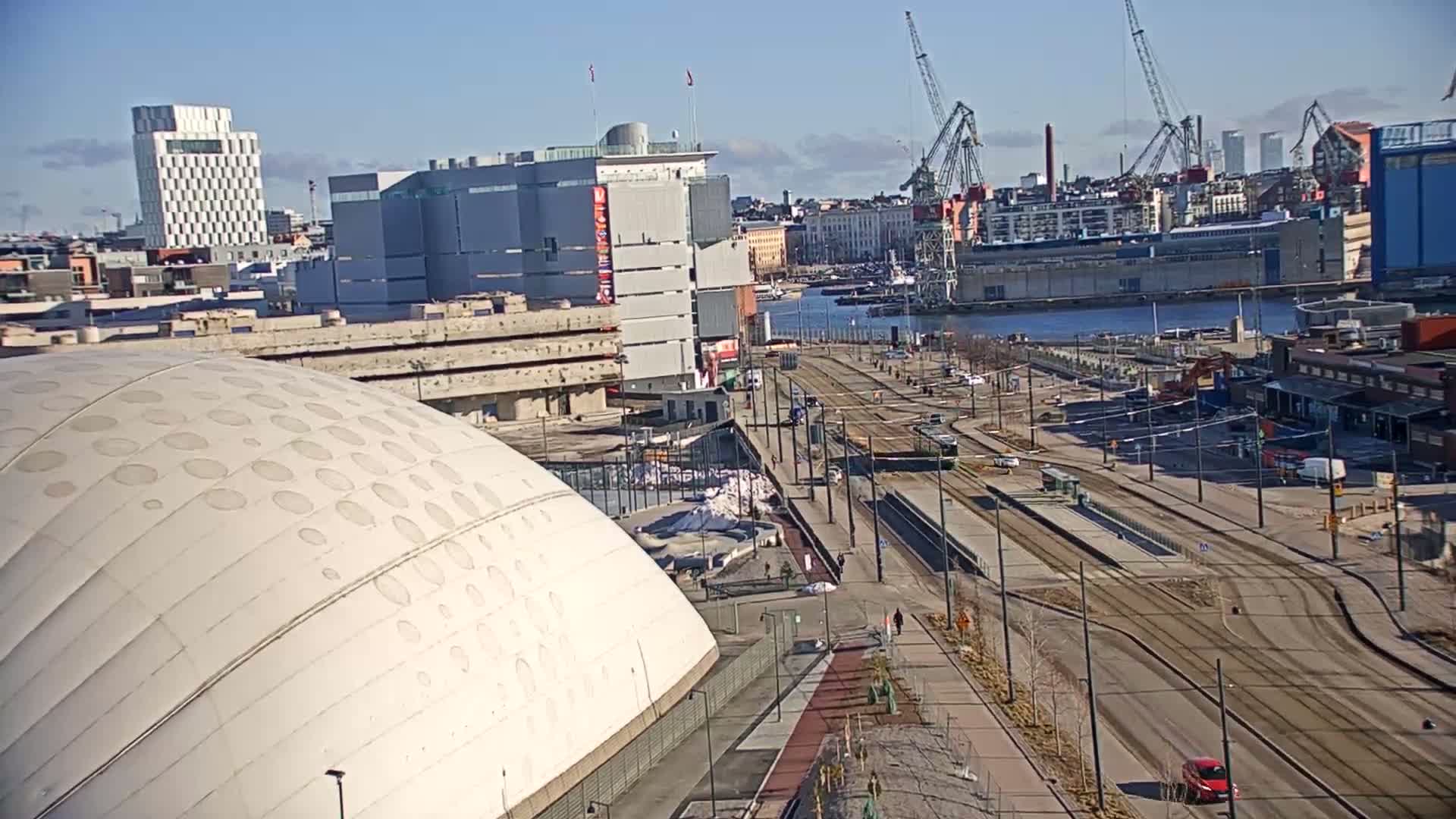 A waterfront city scene on a sunny day shows a large domed building, tram tracks, and several cranes near a harbor.