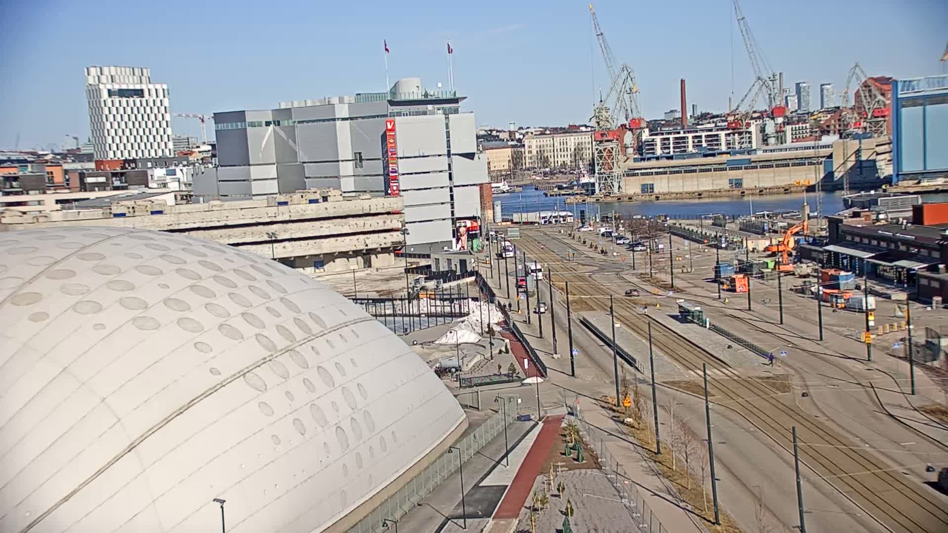 A large domed building is partially visible in the foreground, with a city waterfront, tram tracks, and construction activity in the middle ground, all under a clear sunny sky.
