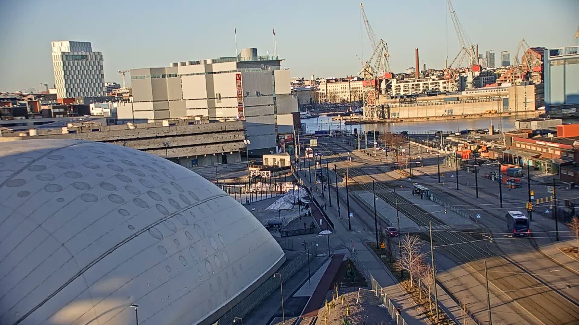 A high-angle, sunny view of a city harbor with a large domed building in the foreground, tram lines, and several cranes in the background.