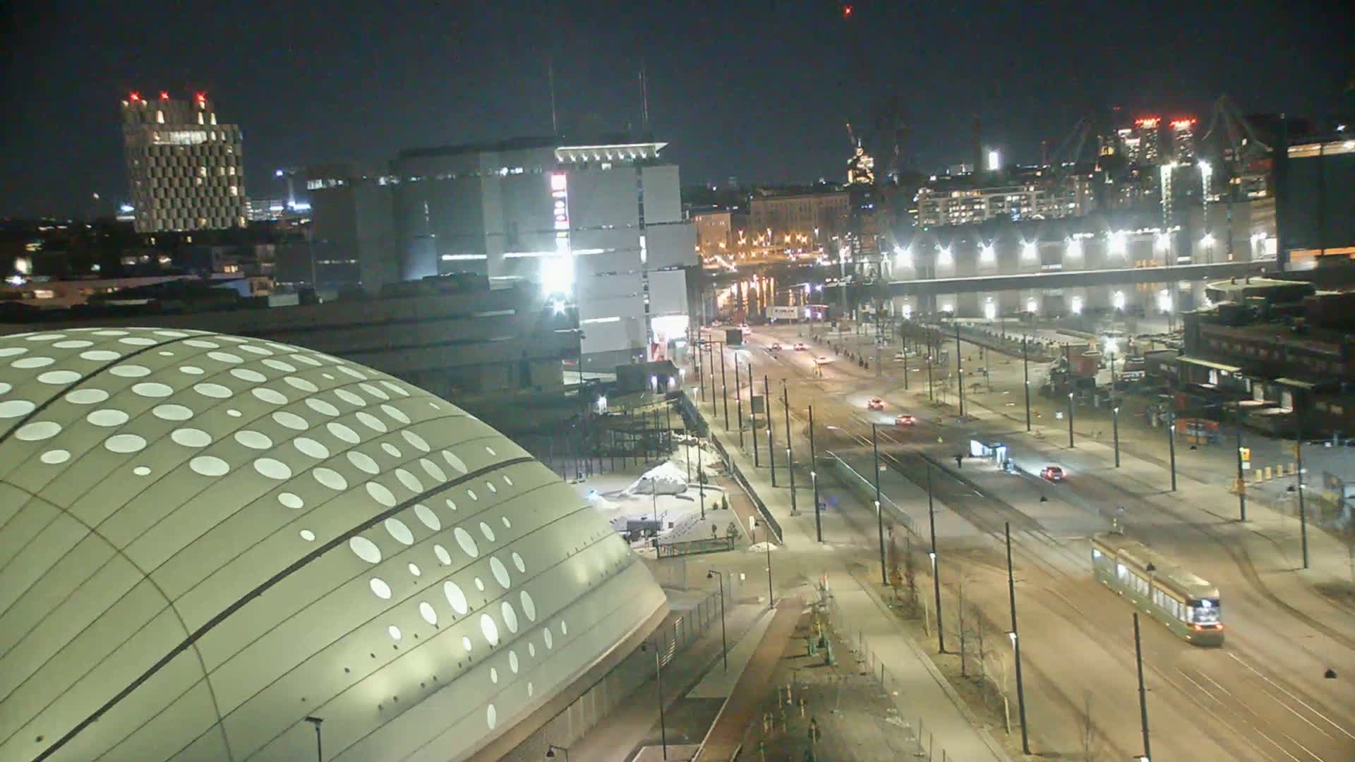 A nighttime, city street scene showing a tram and cars traveling on a wide road past a domed building with white circles on its surface, alongside other buildings and industrial structures.