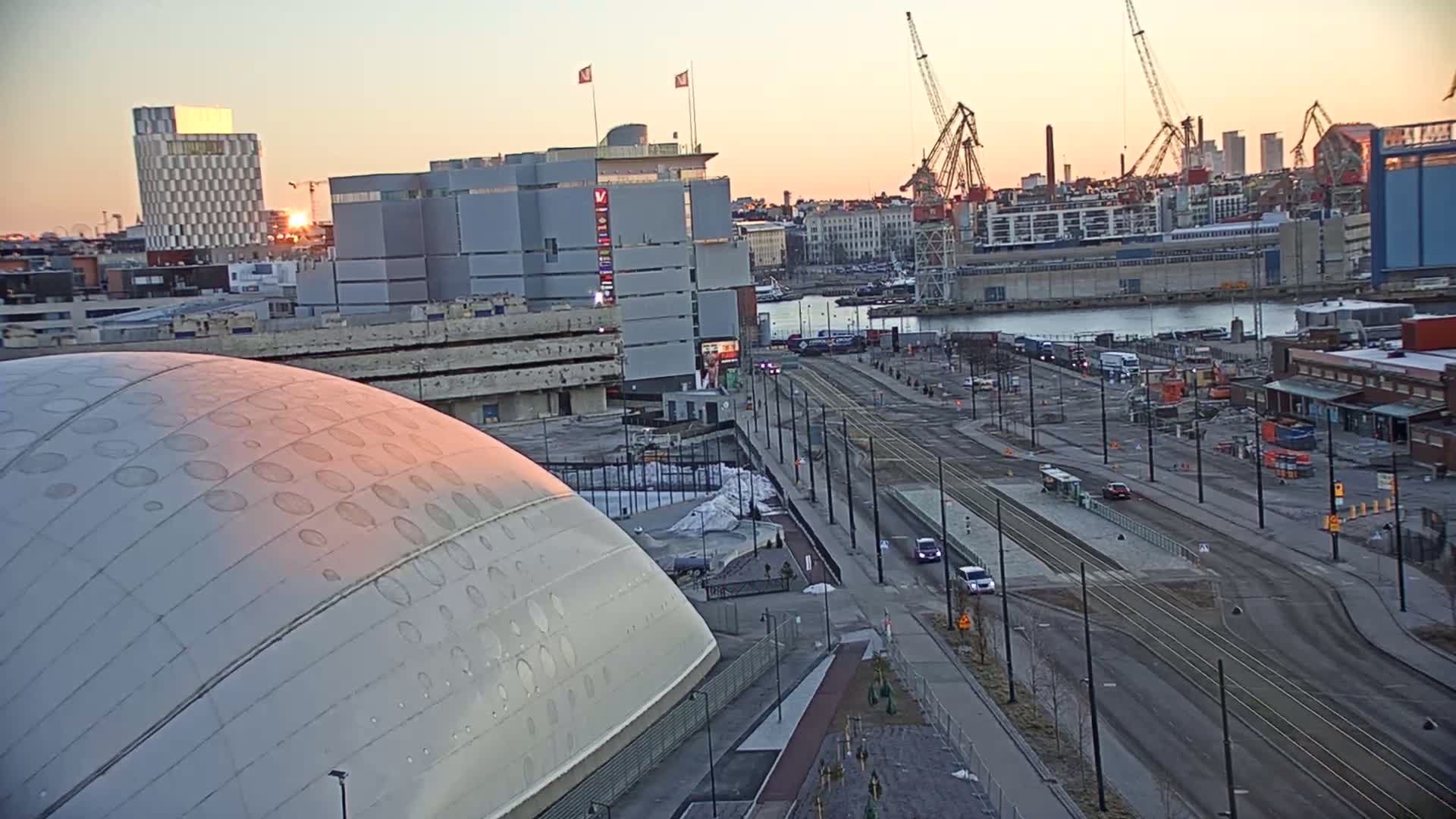 A large domed building is seen at sunset next to a tram line and harbor with several cranes, buildings, and vehicles.