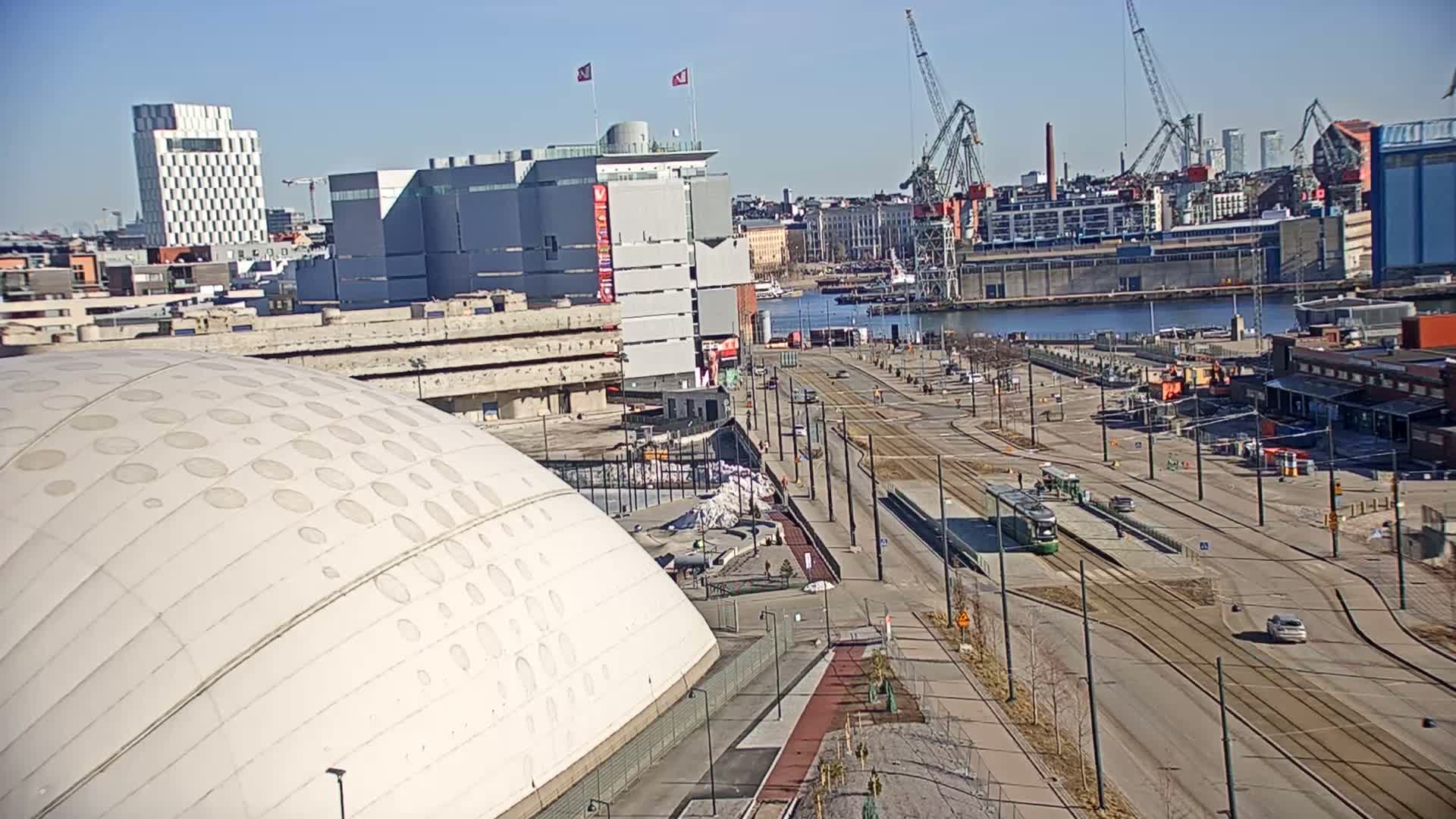 A large, domed white building sits beside a waterfront city street with tram tracks, under a clear blue sky.