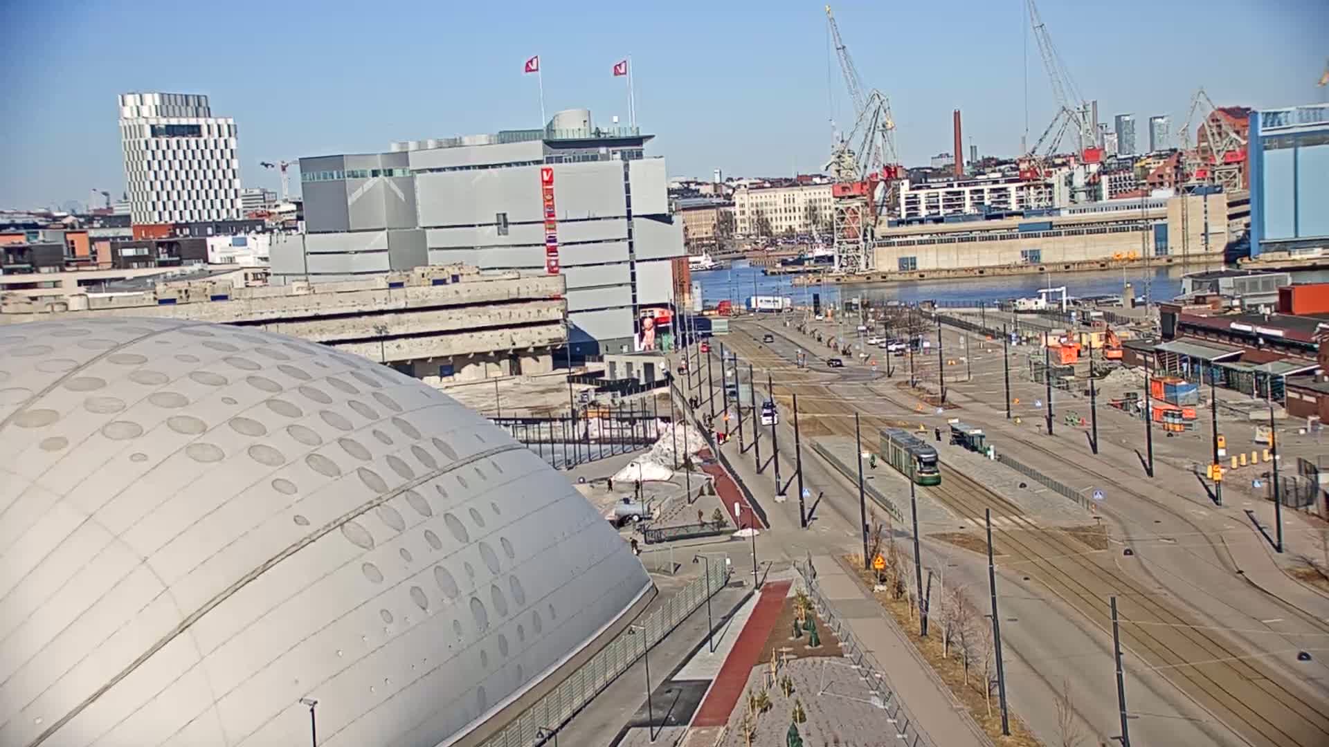 A large domed building sits beside a mostly empty city street with tram tracks, cranes, and a body of water in the background under sunny skies.