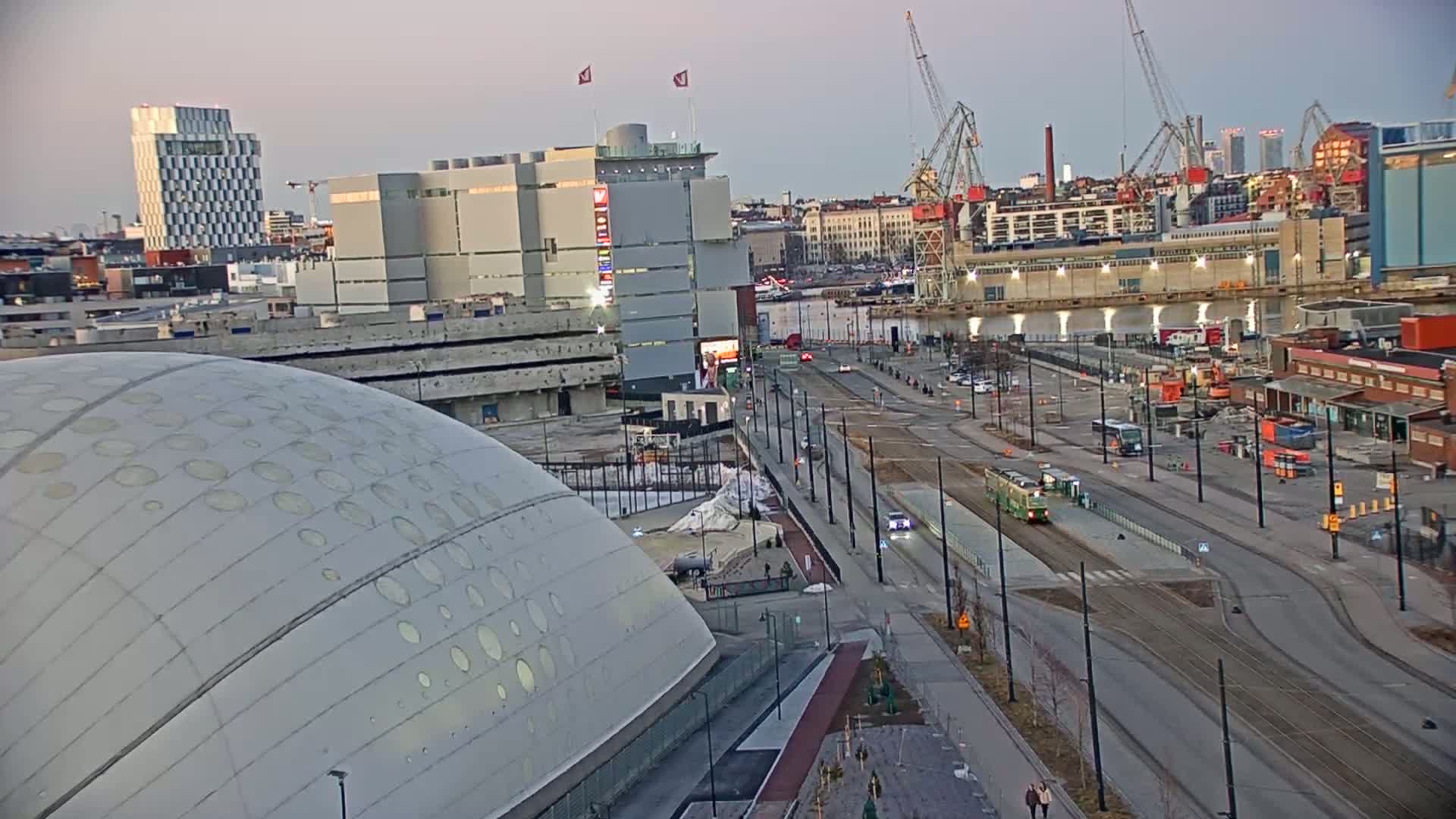 A large, domed building with patterned holes is adjacent to a city street with a tram, construction sites, and waterfront industrial areas under a twilight sky.