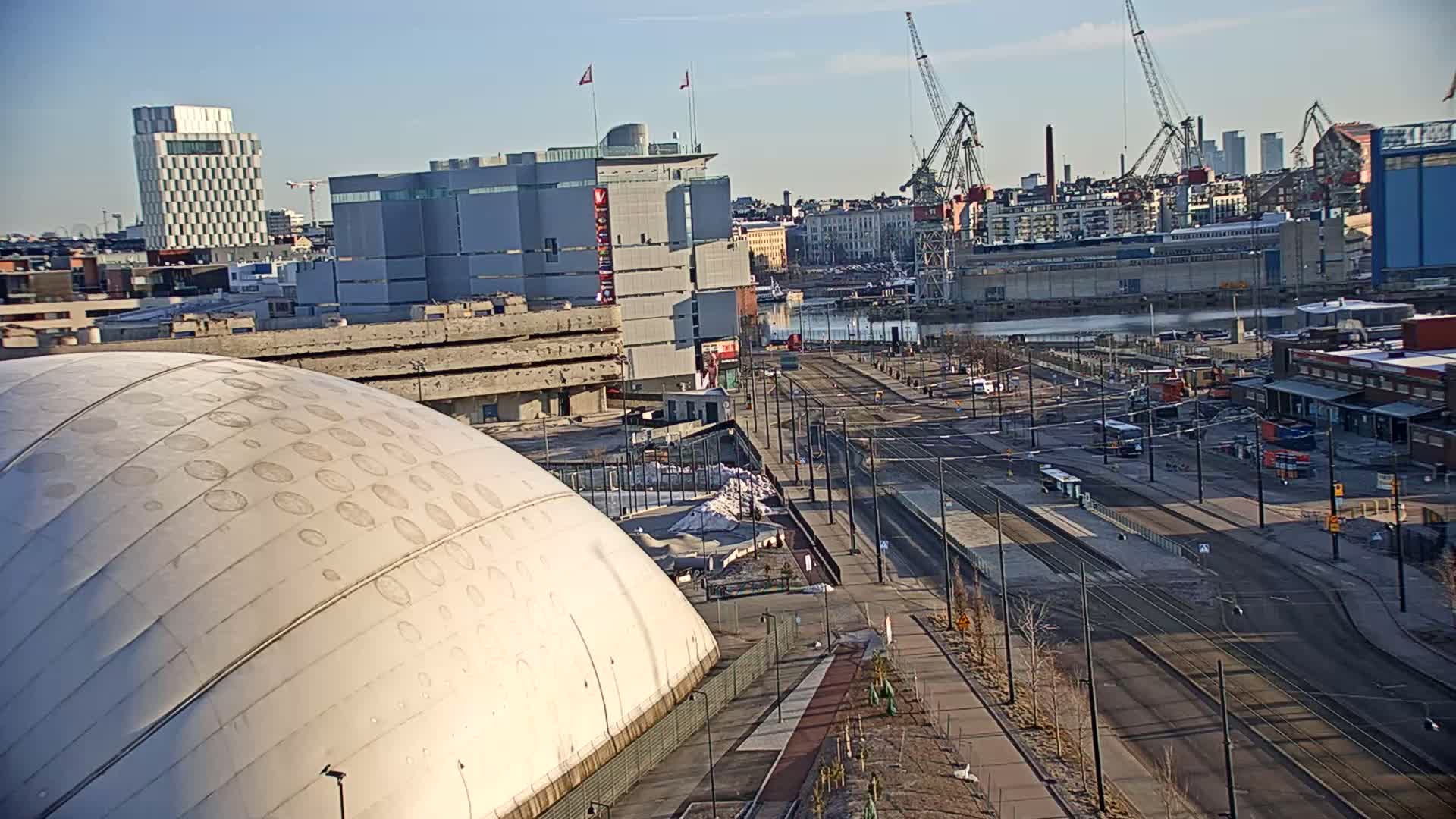 A large, domed structure is situated near a waterfront area with urban buildings, tram tracks, and several cranes under a clear, sunny sky.