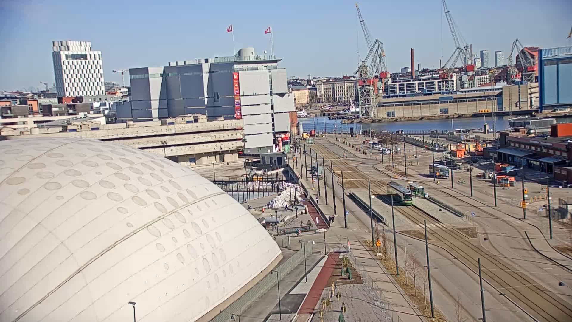 A high-angle view on a sunny day shows a large domed building, tram tracks, a waterfront with cranes and industrial buildings, and several other buildings in the background.