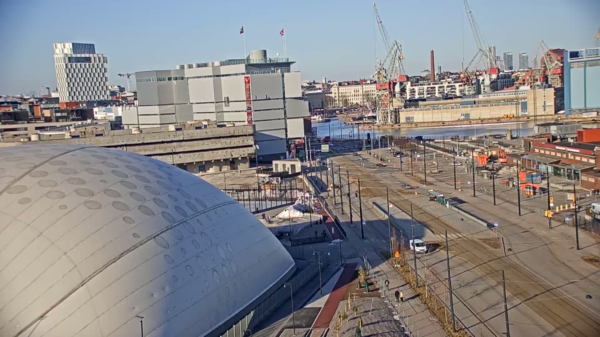 A large, domed building sits near a body of water with several cranes and urban structures visible in the background on a sunny day.