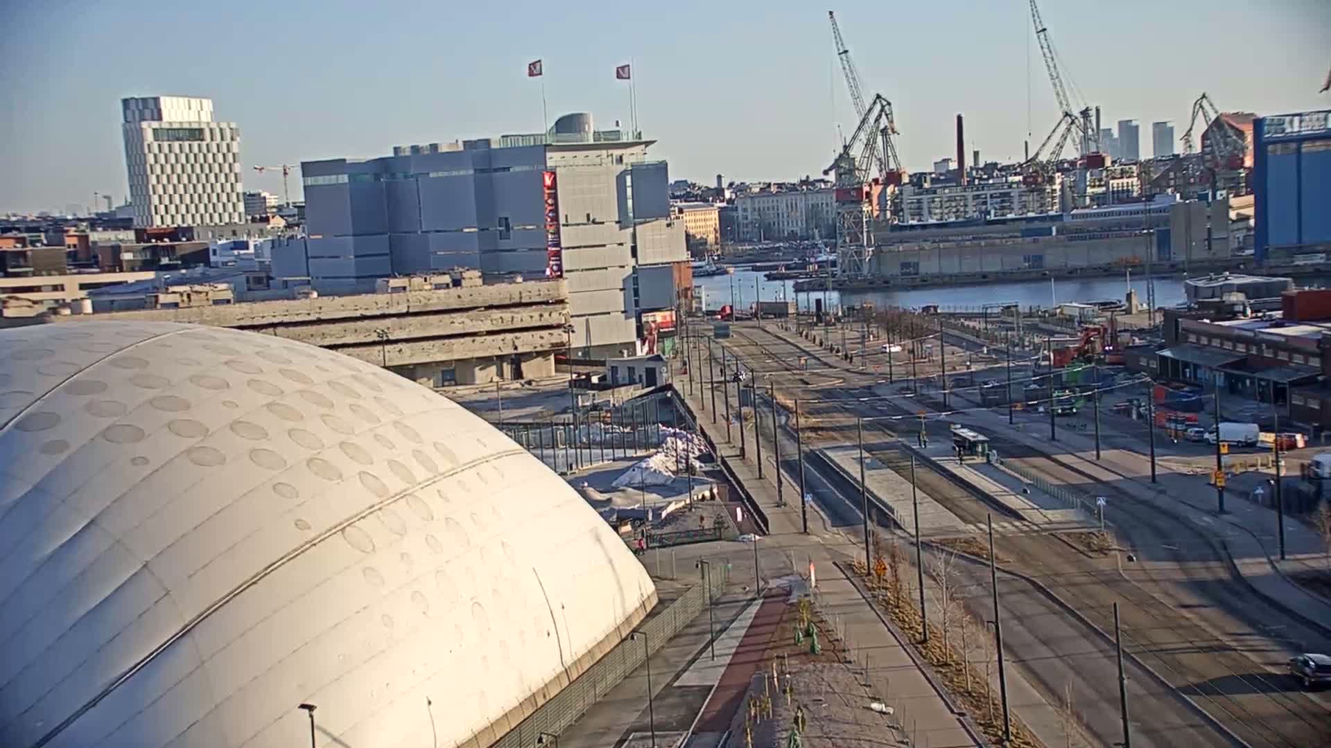 A high-angle view shows a cityscape on a sunny day, featuring a large domed building in the foreground, roads and tram lines leading to a harbor with cranes and ships, and various buildings in the background.