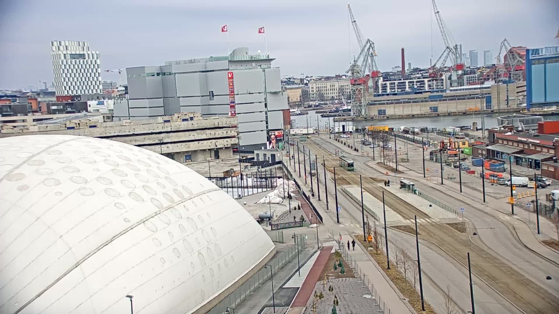 A large white dome-shaped building sits beside a wide, mostly empty road with tram tracks, under a cloudy sky, overlooking a harbor with cranes and various buildings.
