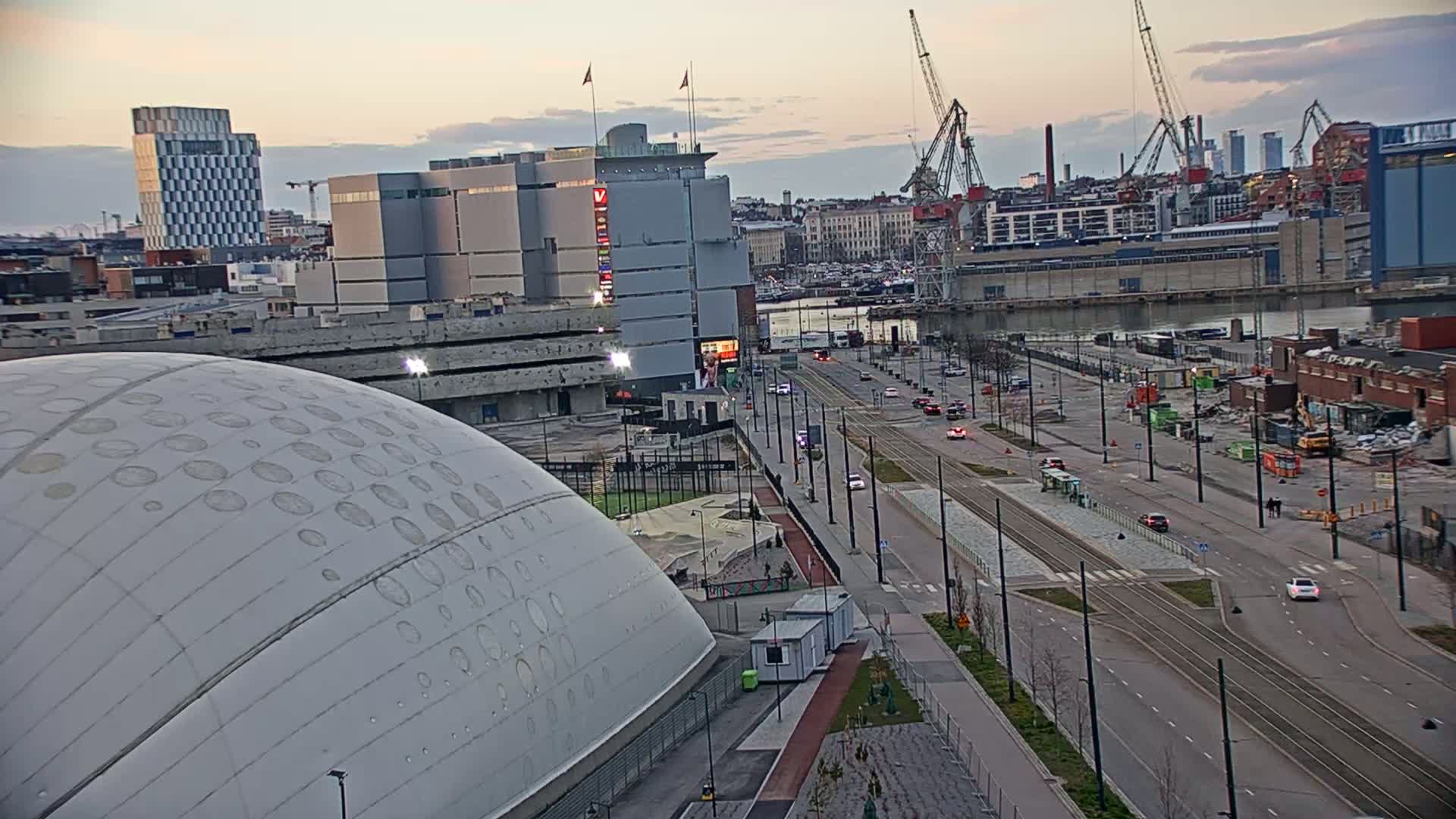 A large, domed building sits near a waterfront city street with tram tracks, under a twilight sky.