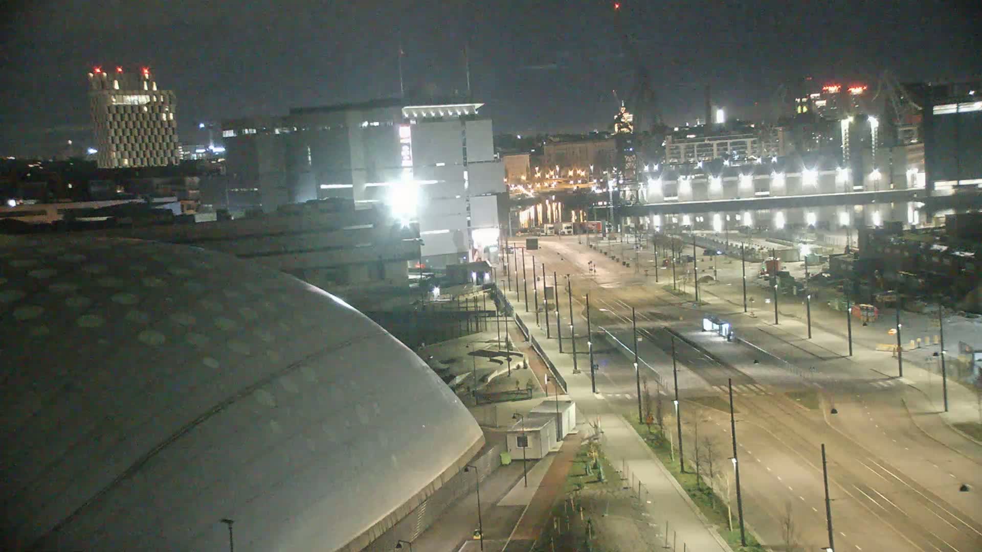 A nighttime, city view shows a large domed building in the foreground, empty streets and tram tracks, and various other buildings and structures in the background under a dark sky.