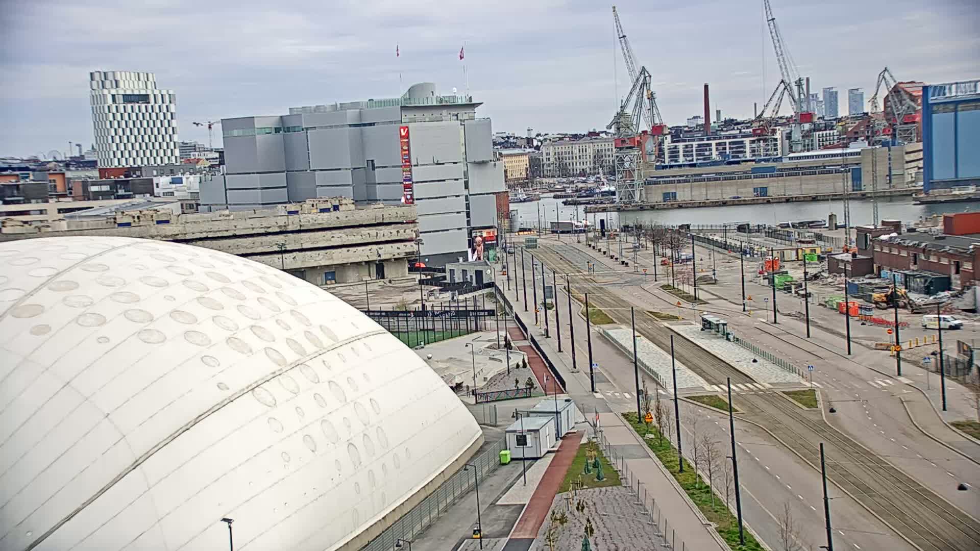A large white domed building sits near a wide, mostly empty road and tram tracks, with a harbor and city skyline visible in the background under a cloudy sky.