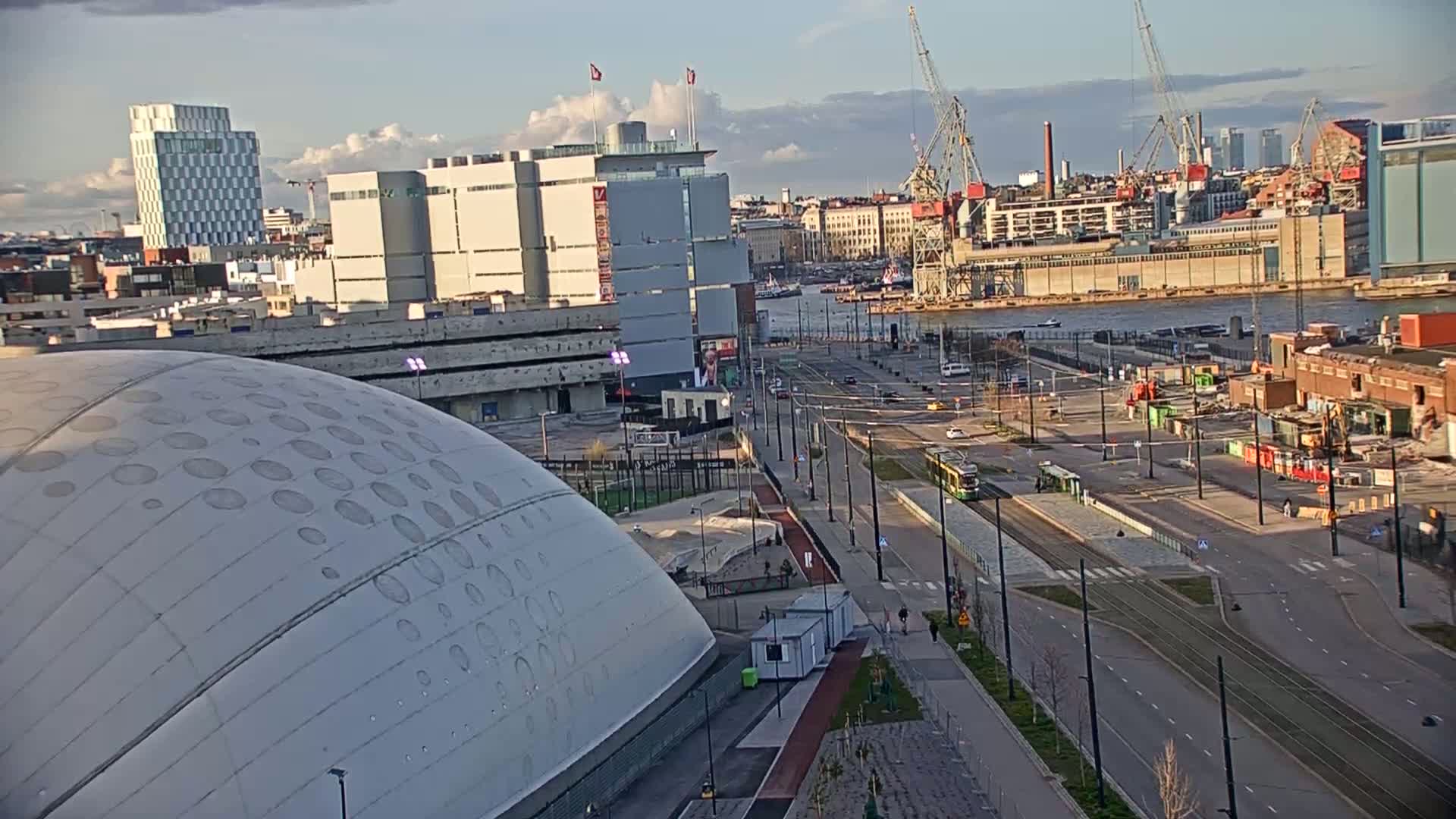 A mostly cloudy, late afternoon scene overlooks a city with a large domed building in the foreground, streets, trams, and a harbor with cranes in the background.