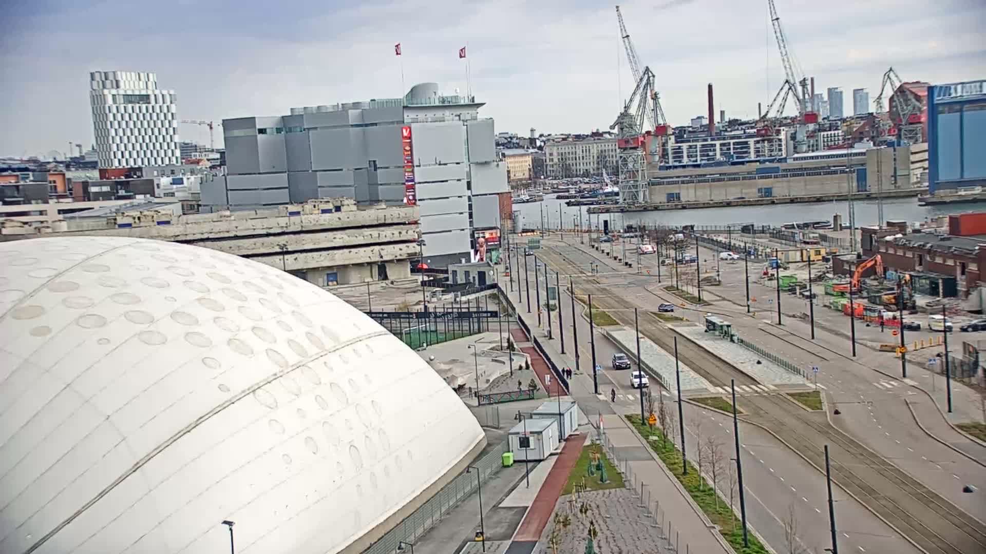 A large, domed white building sits next to a city street with tram tracks, overlooking a harbor with cranes and other industrial structures under a partly cloudy sky.