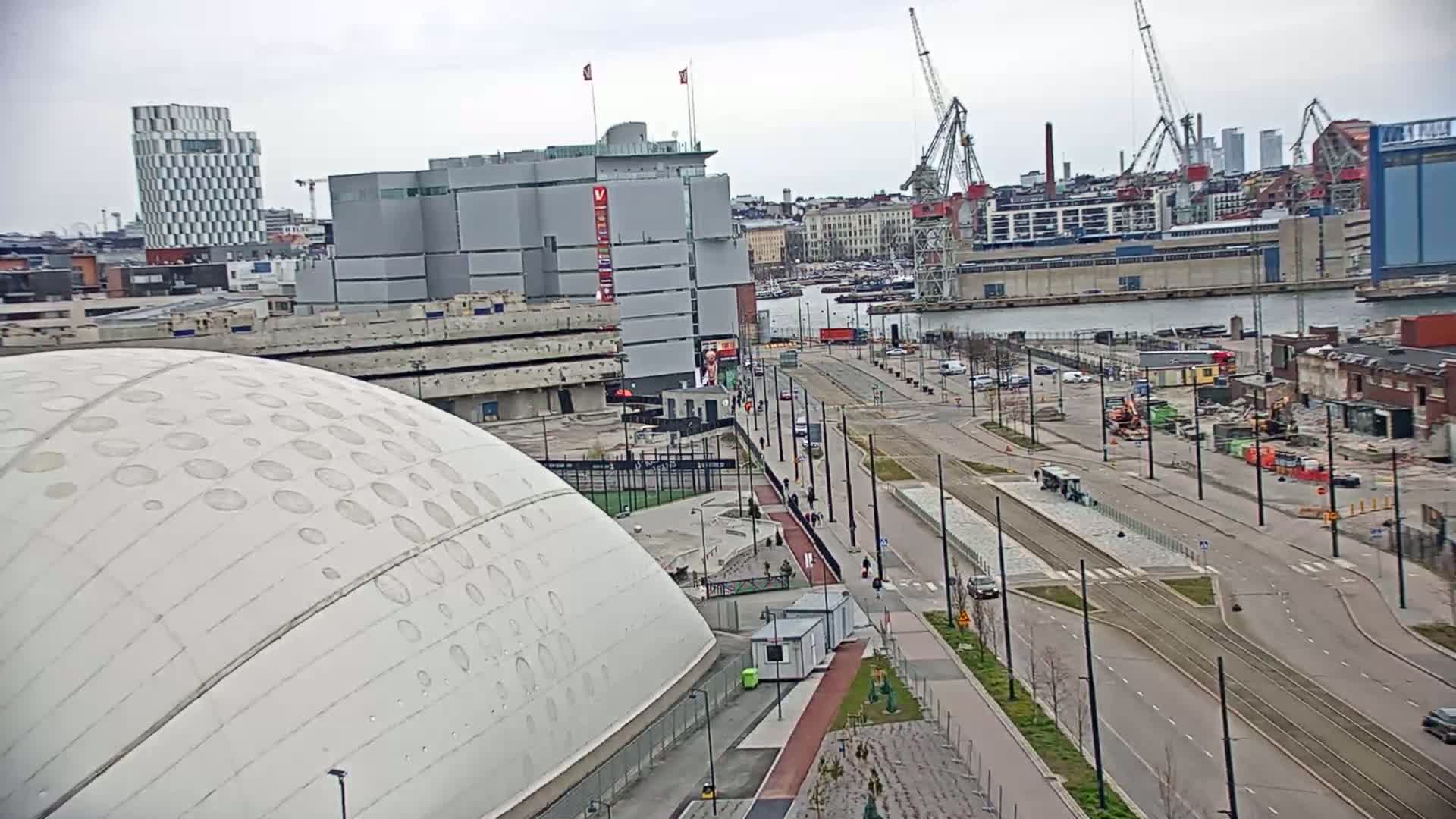 A large, domed building with patterned circles sits next to a wide, mostly empty street with tram tracks, near a waterfront with construction cranes and docked ships under an overcast sky.