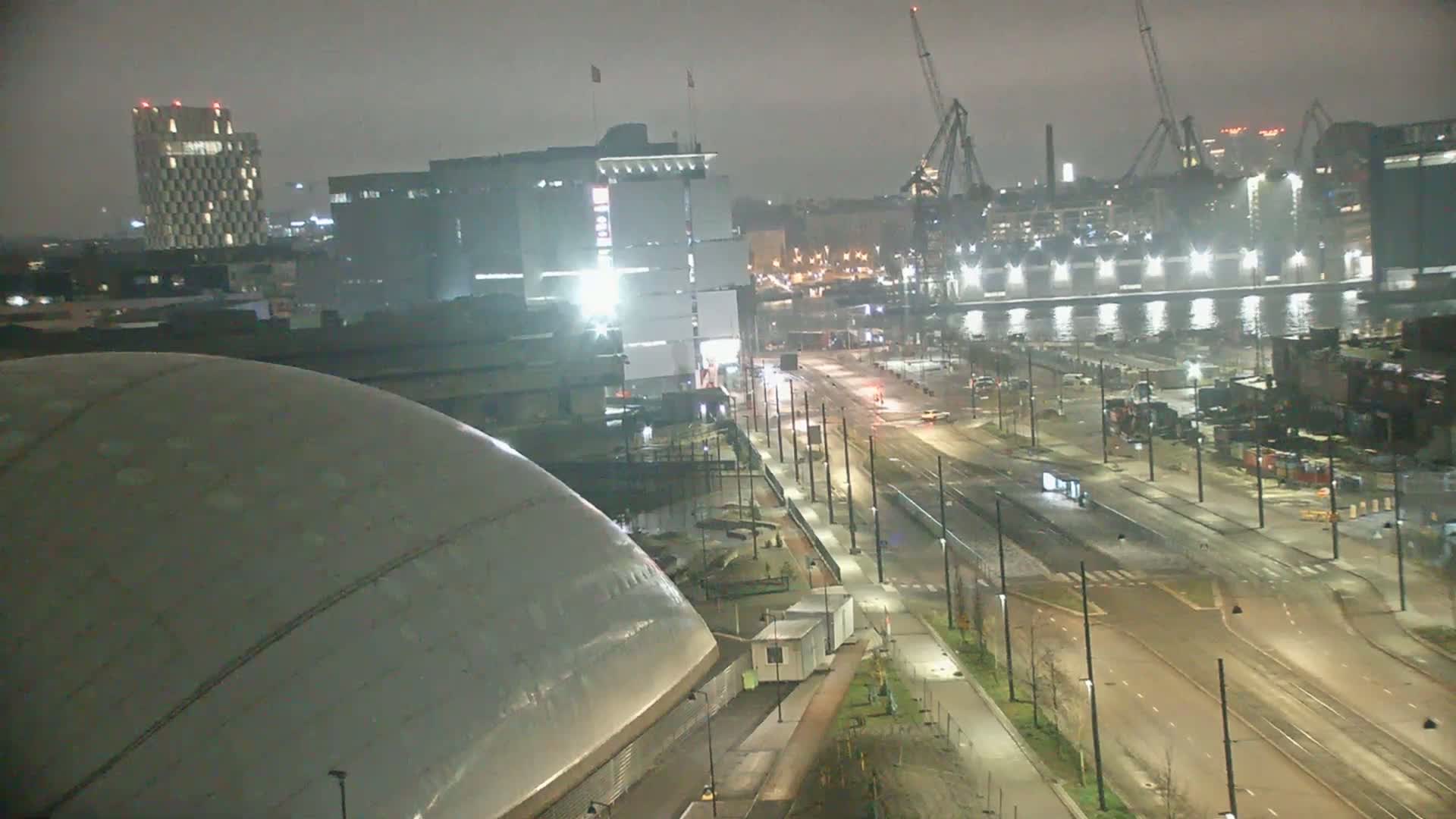 A nighttime, overcast view of a city harbor with a large, domed building in the foreground, a multi-lane road with tram lines in the middle ground, and several buildings and industrial structures in the background.