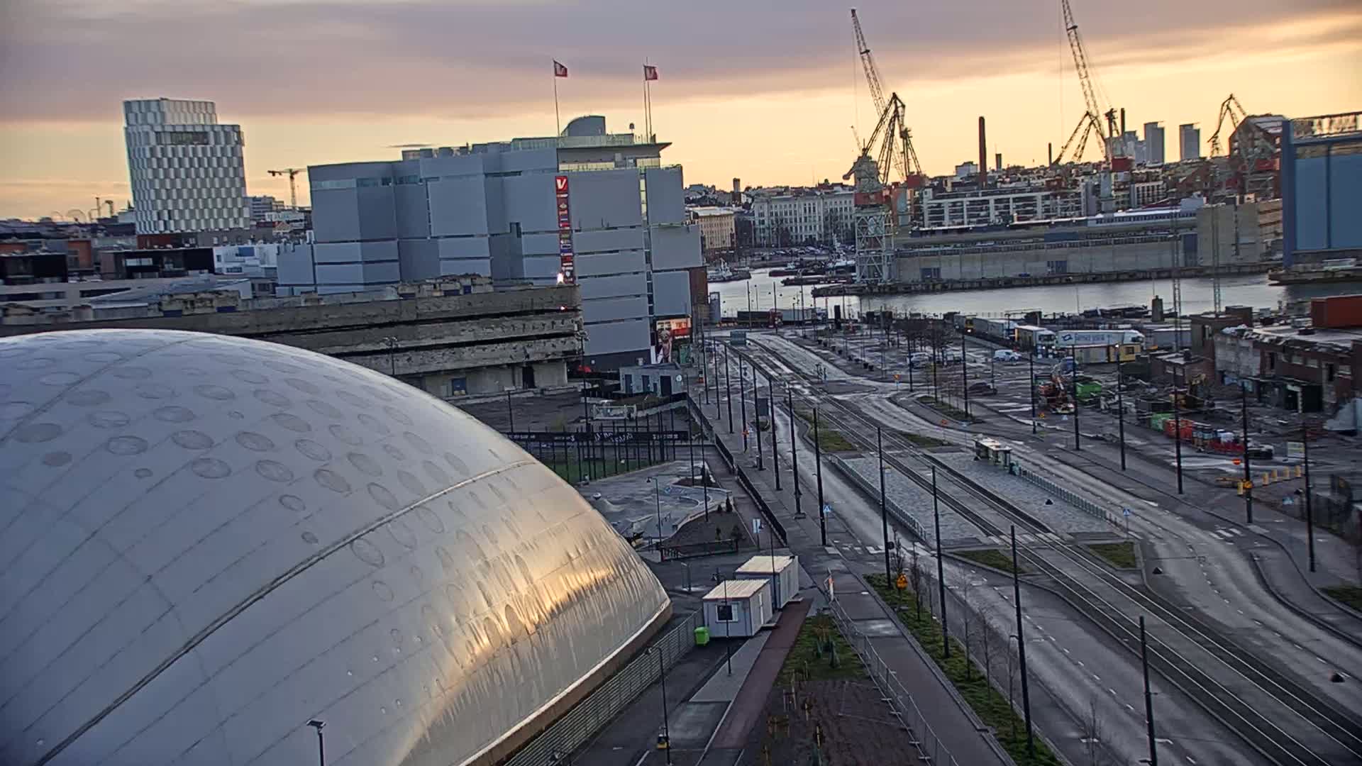 A large, dome-shaped building is situated near a wide road with tram tracks, overlooking a harbor filled with ships and cranes under a dusky sky.