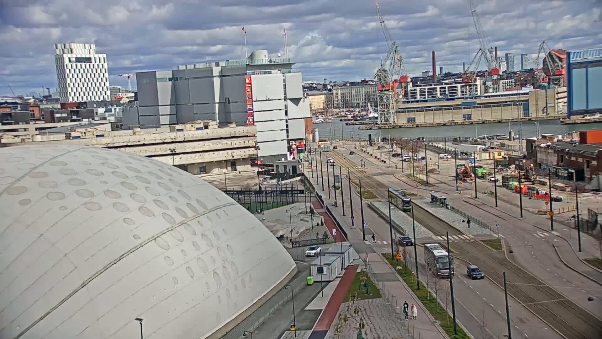 A partly cloudy day reveals a city waterfront scene with a large domed building, modern architecture, trams on a roadway, and construction activity near a harbor.