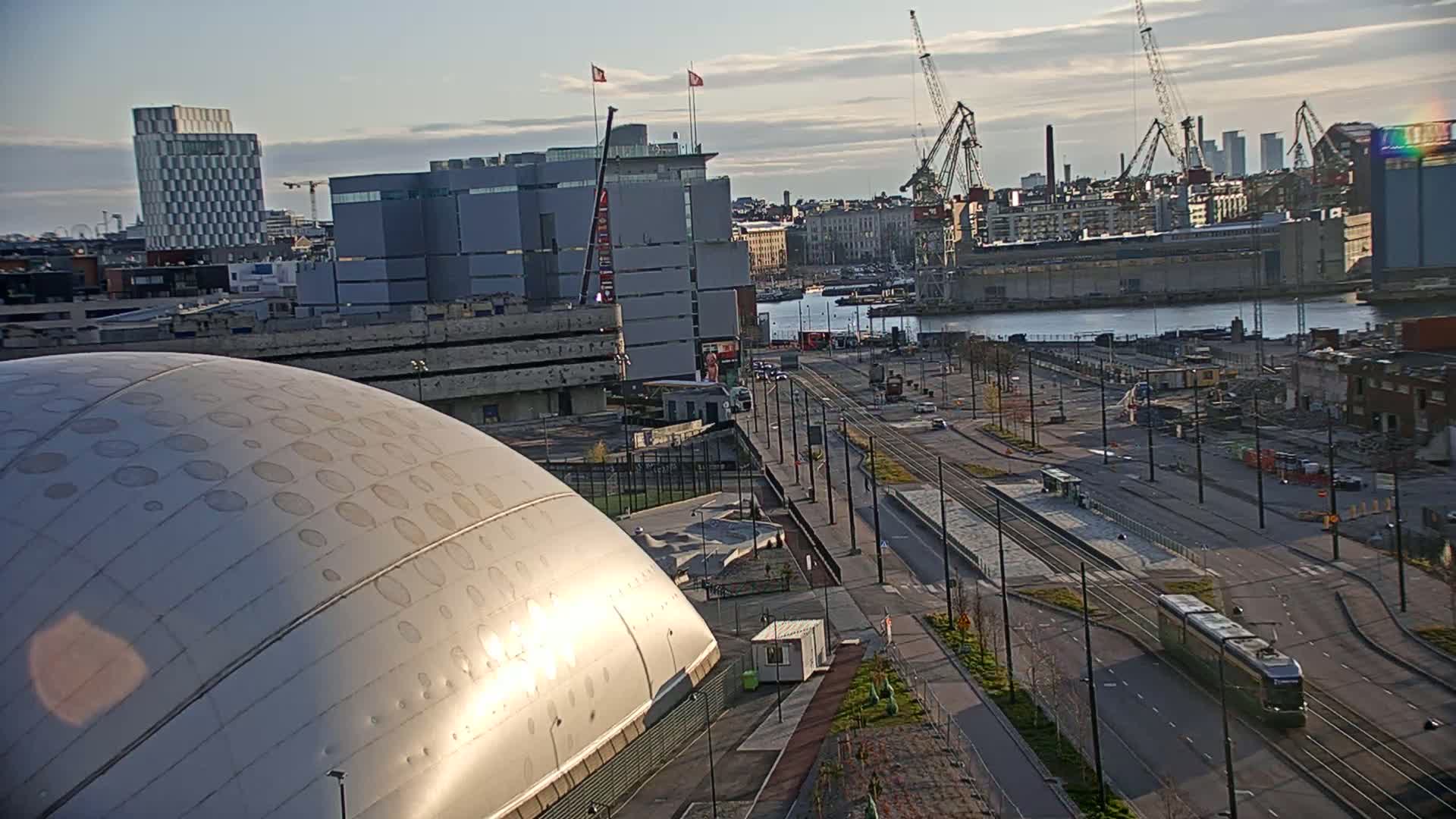 A large, metallic dome-shaped building sits in the foreground, overlooking a city scene with a tram and harbor under a partly cloudy sky.