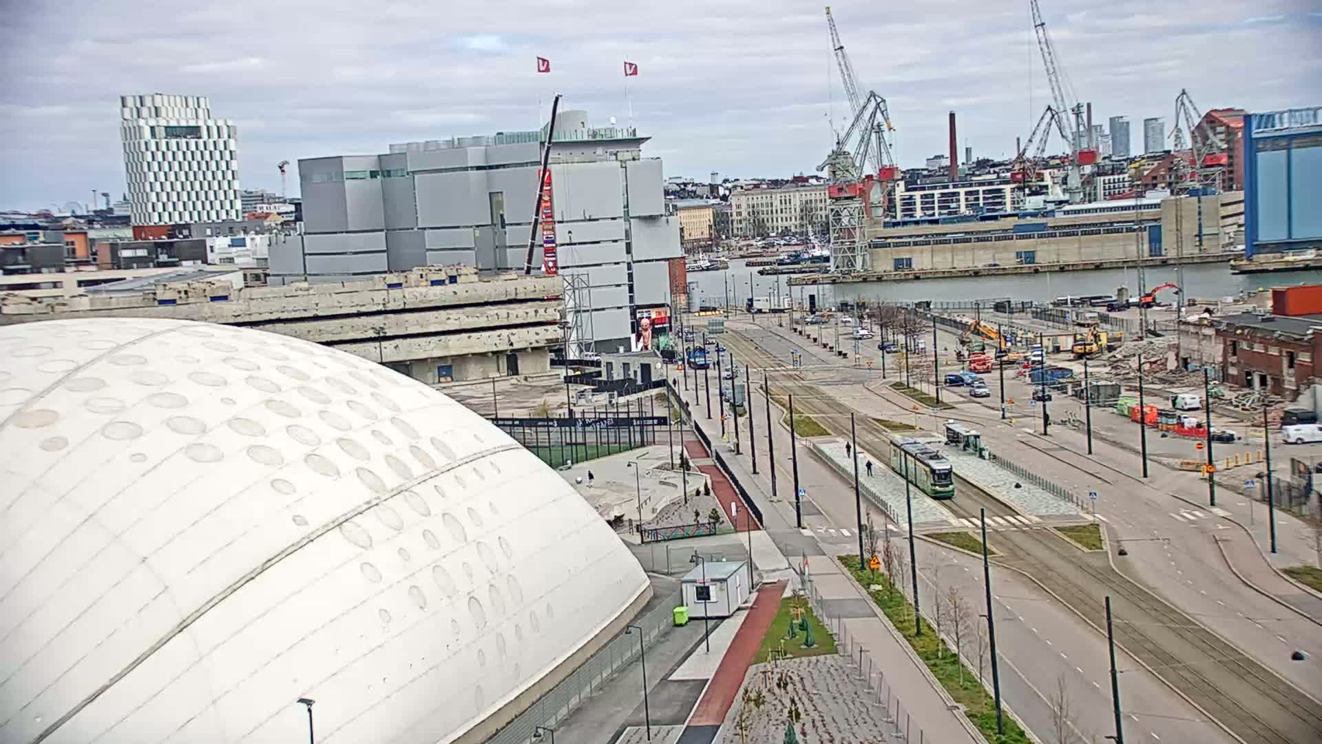 A large domed building is situated near a waterfront city scene with overcast skies, construction equipment, and a tram on a wide street.
