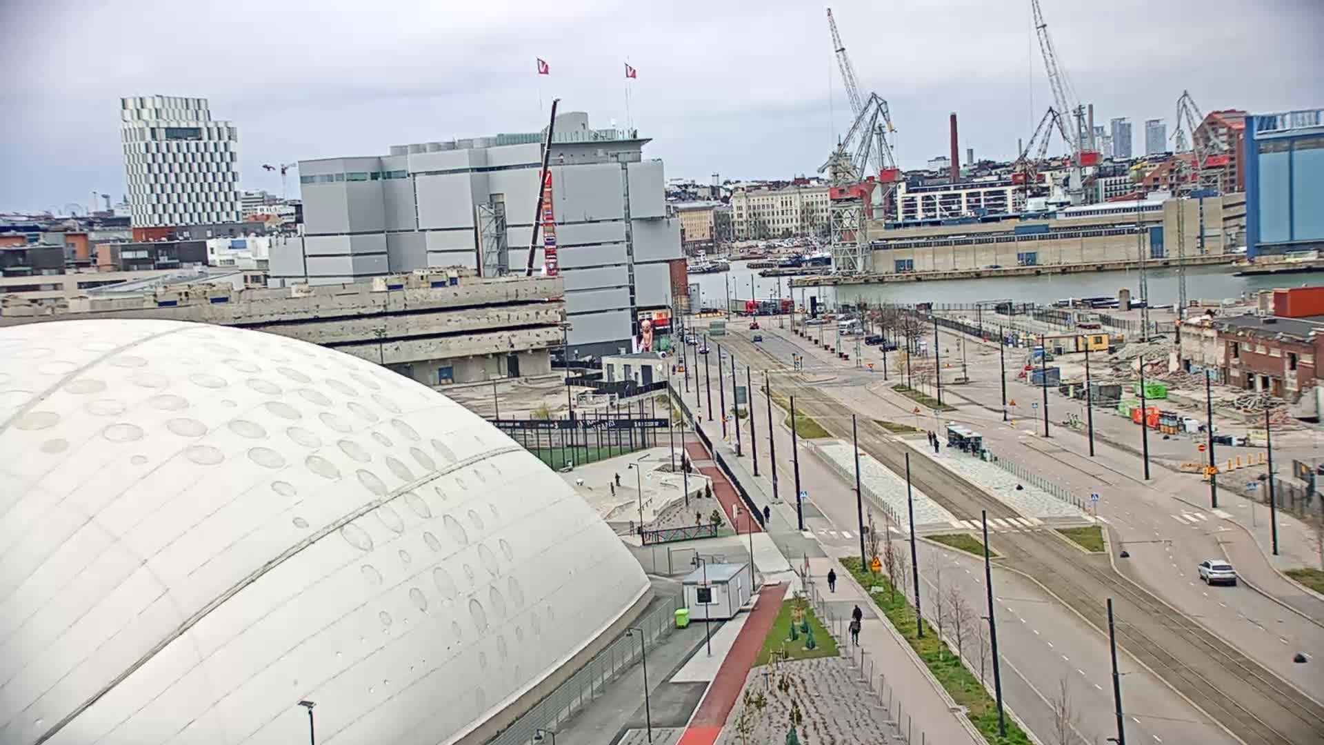 A mostly cloudy day reveals an urban waterfront scene with a large domed building, roads, construction sites, and several cranes at a shipyard.