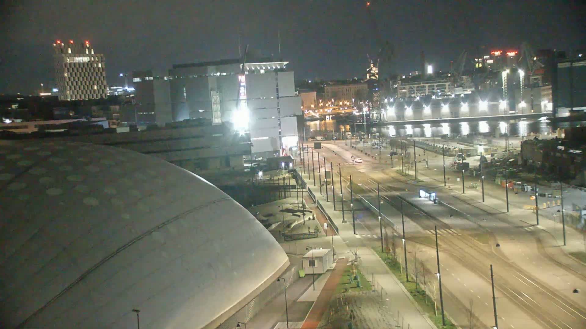 A nighttime aerial view shows a city street with tram tracks, lined with buildings including a large domed structure and a tall rectangular one, under a dark sky.