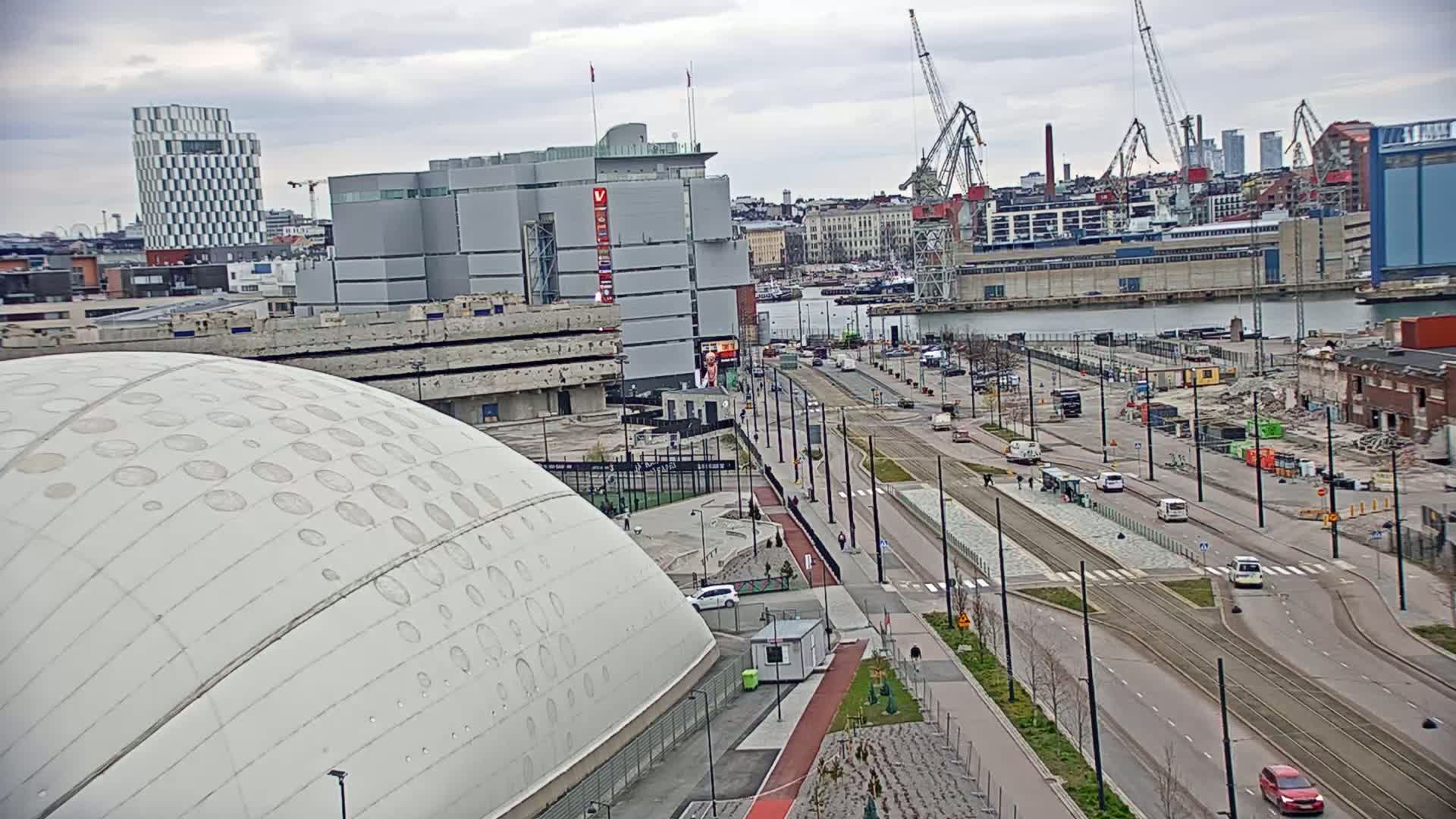 A large, domed building is situated near a city harbor under an overcast sky, with a wide road and tram tracks running past construction sites and cranes in the background.