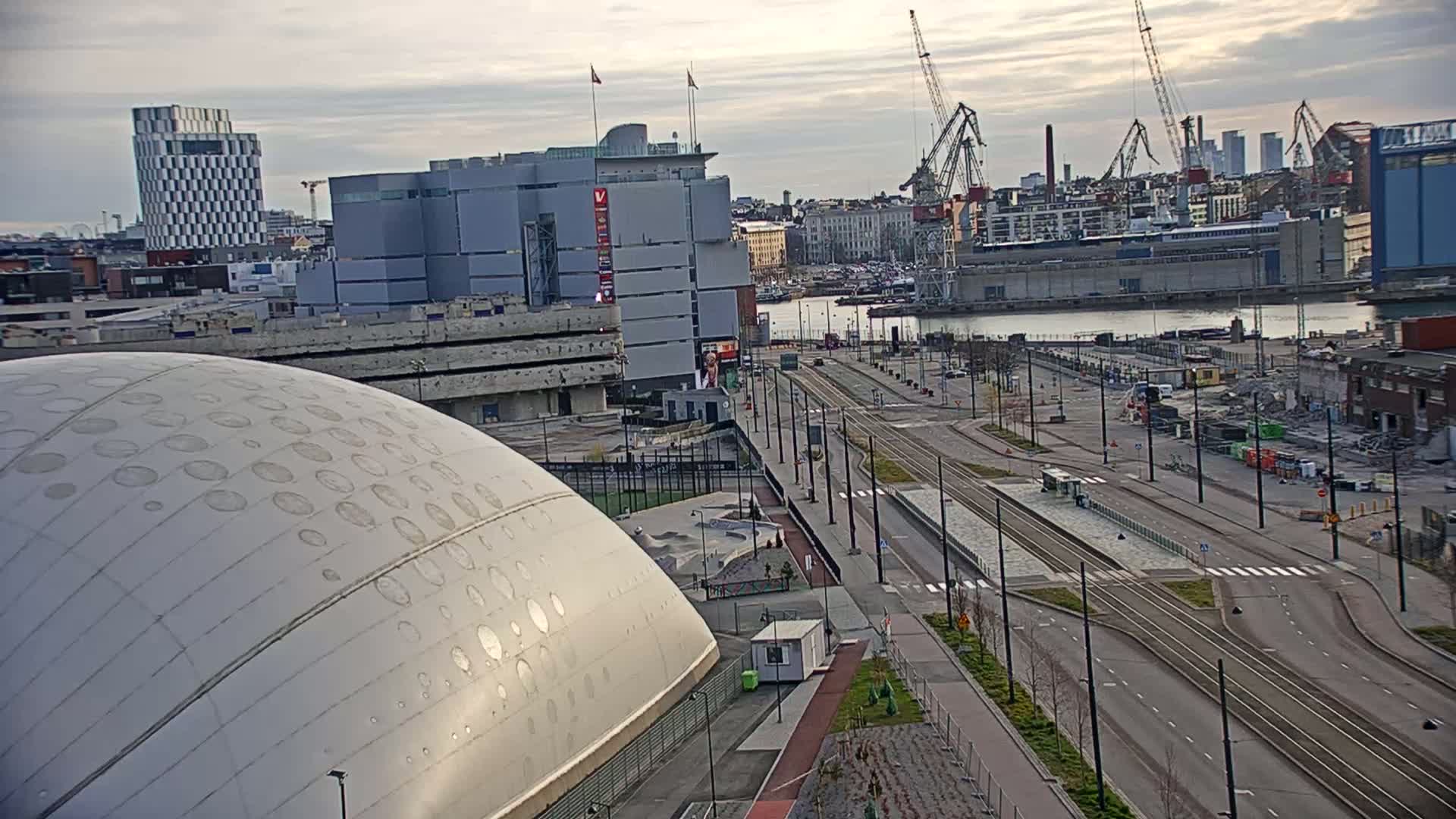 A large, domed building sits next to a wide road with tram tracks running through it, under a partly cloudy sky, with a harbor and cranes visible in the background.