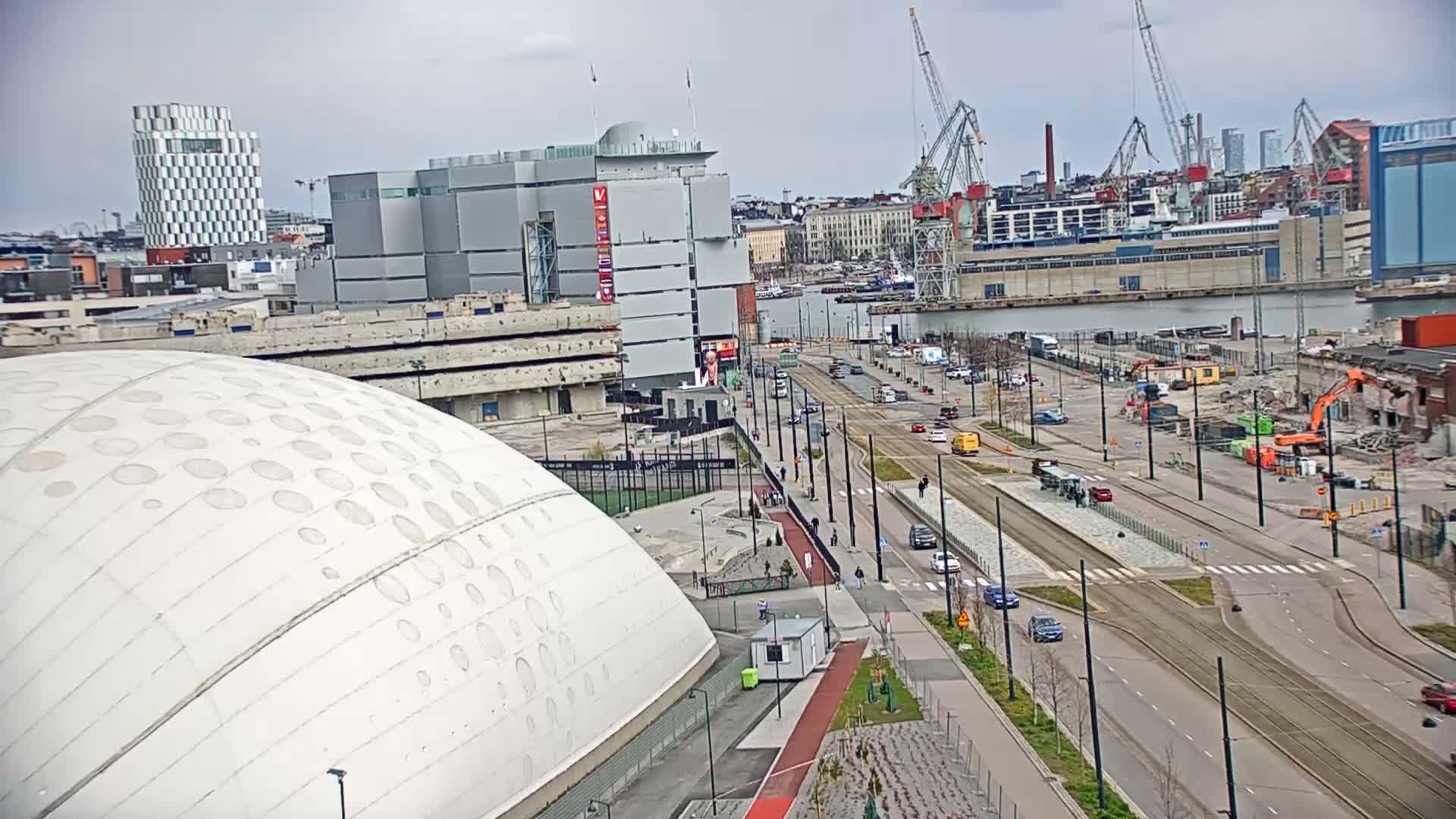 A large white domed building is situated near a waterfront city street with tram tracks, under a cloudy sky, with construction and demolition visible in the distance.
