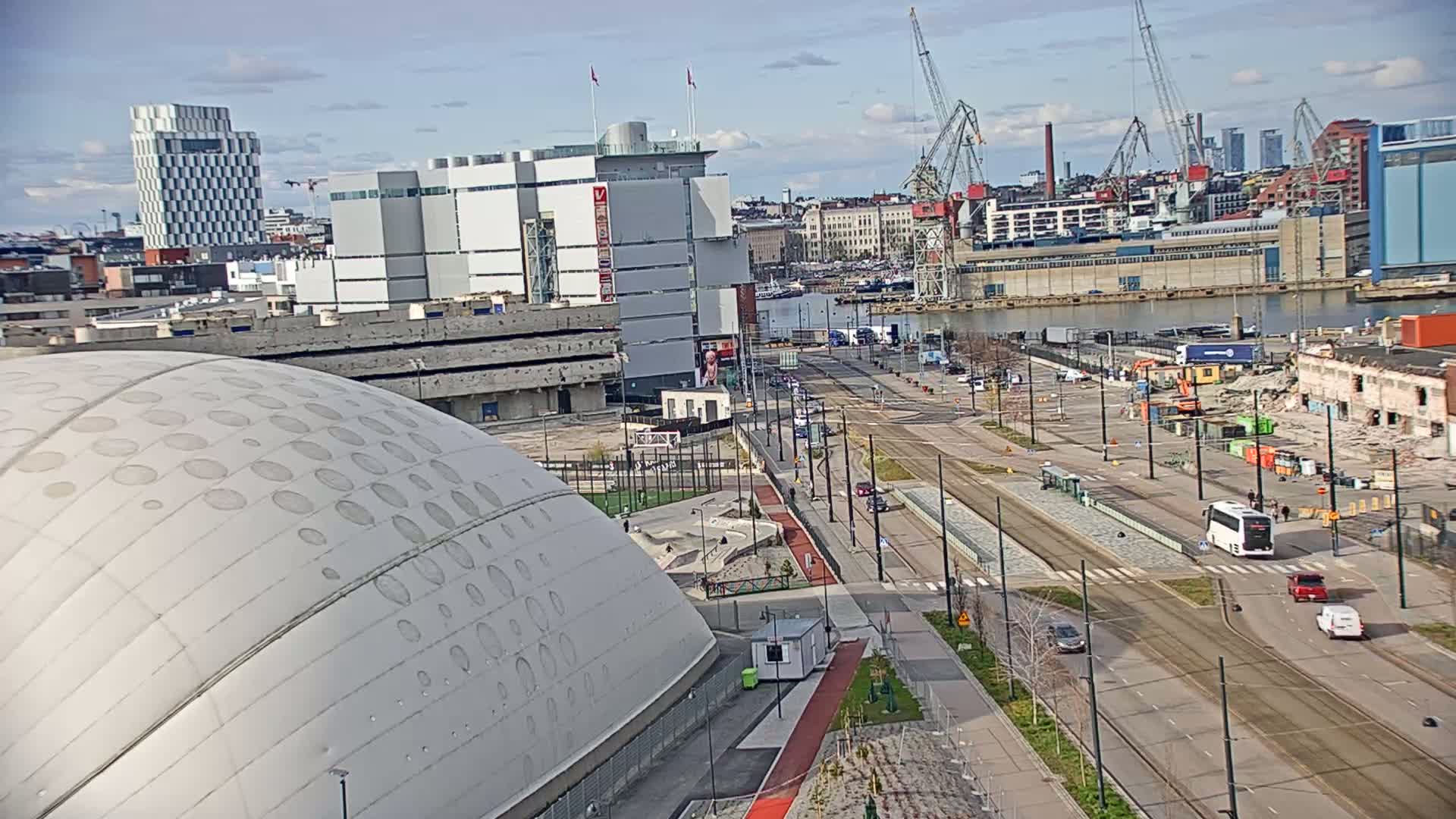 A large domed building sits beside a city street with tram lines, alongside a harbor with industrial buildings and cranes under a partly cloudy sky.