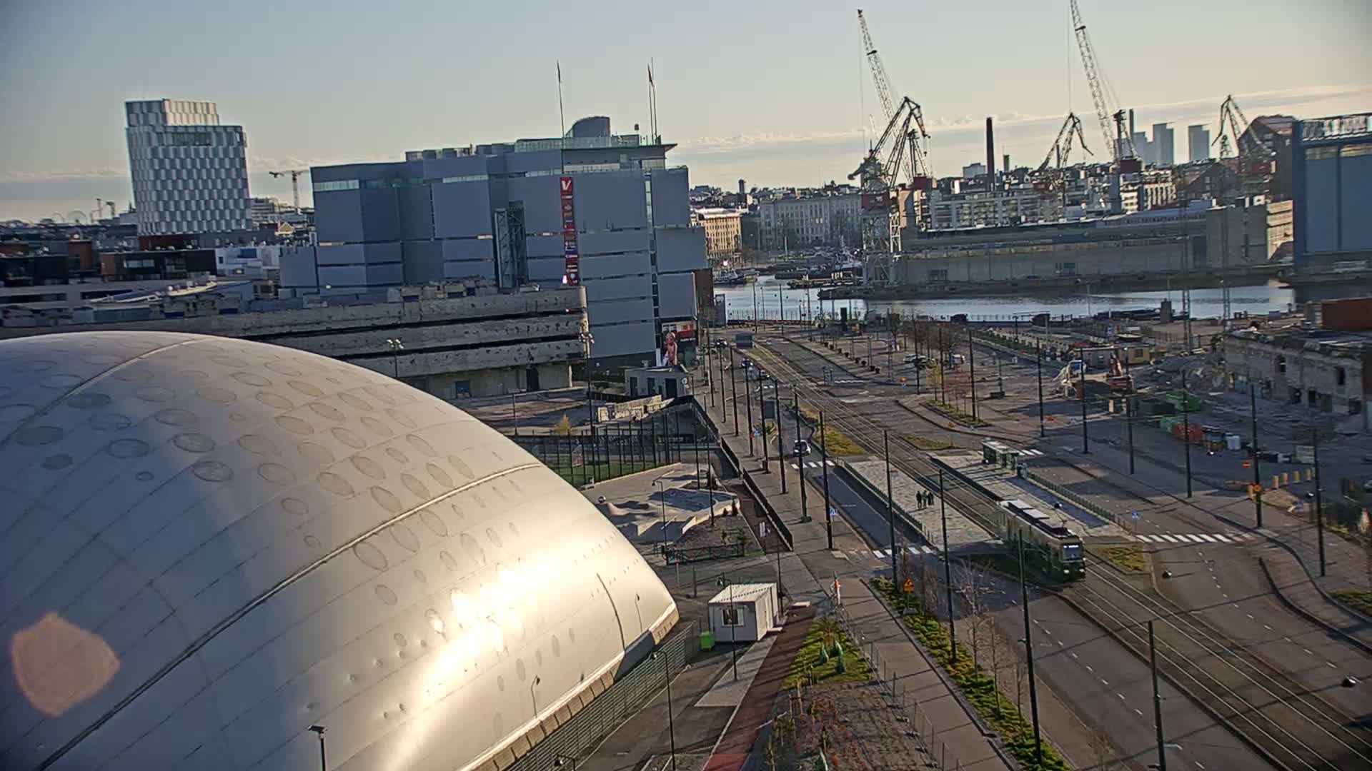 A large, domed building is partially visible in the foreground, with a city landscape including a waterfront, cranes, and a tram in the background under a clear, sunny sky.