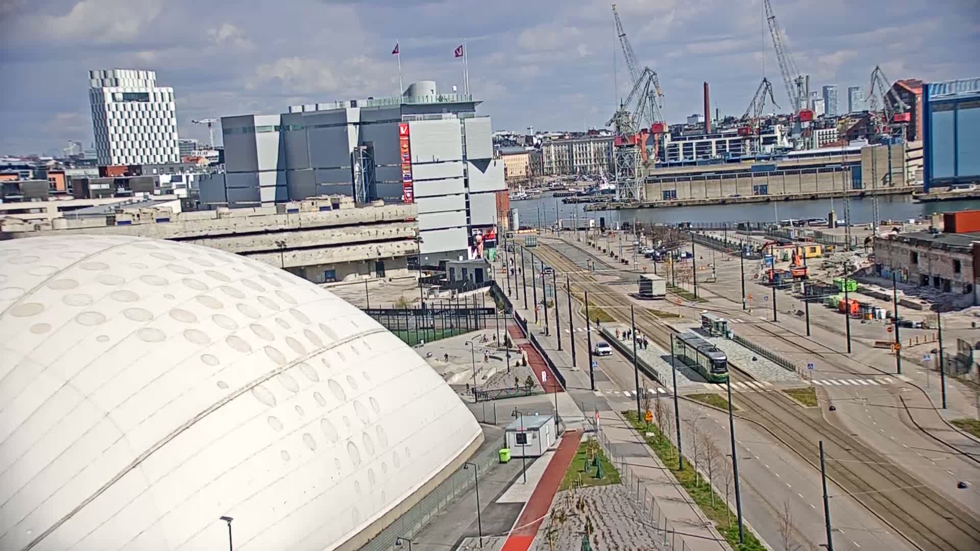 A large, domed white building sits near a waterfront city with tram lines, cranes, and partially demolished structures under a partly cloudy sky.