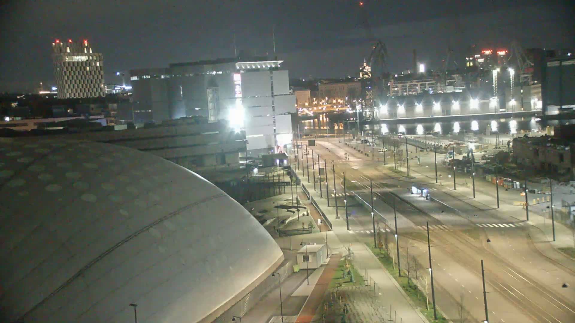 A nighttime view of a city harbor area, showing a large domed building, several other buildings, and a mostly empty roadway with tram tracks.