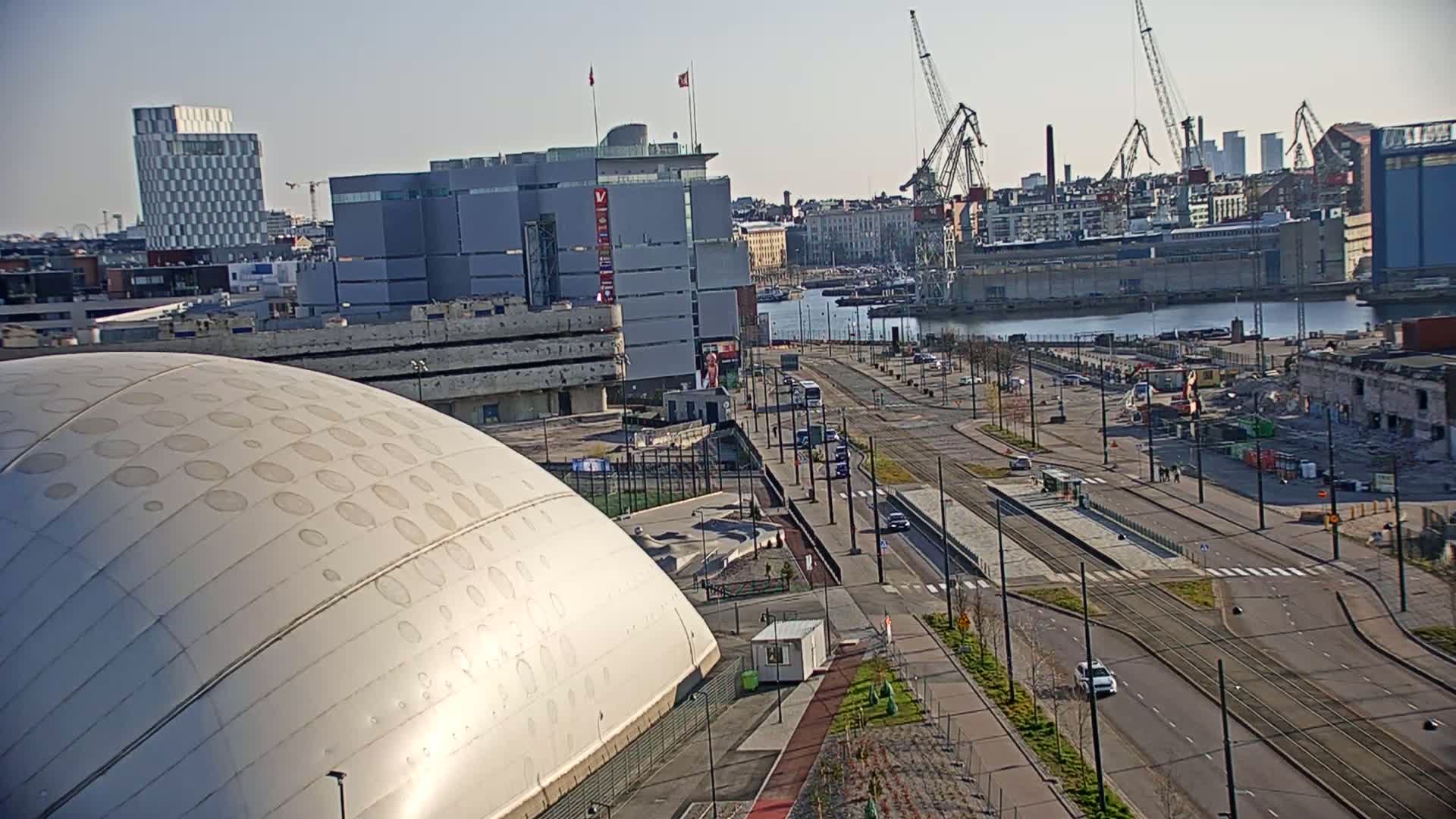 A large, domed building with circular patterns sits in the foreground, next to a roadway with tram tracks, leading to a harbor with several cranes under a clear, sunny sky.