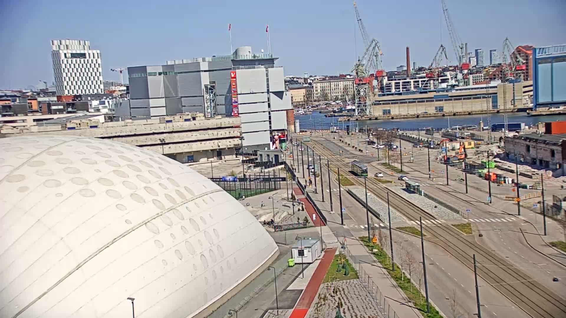 A large, domed building with circular indentations is situated near a waterfront area with tram tracks, sparse traffic, and construction activity under a clear, sunny sky.