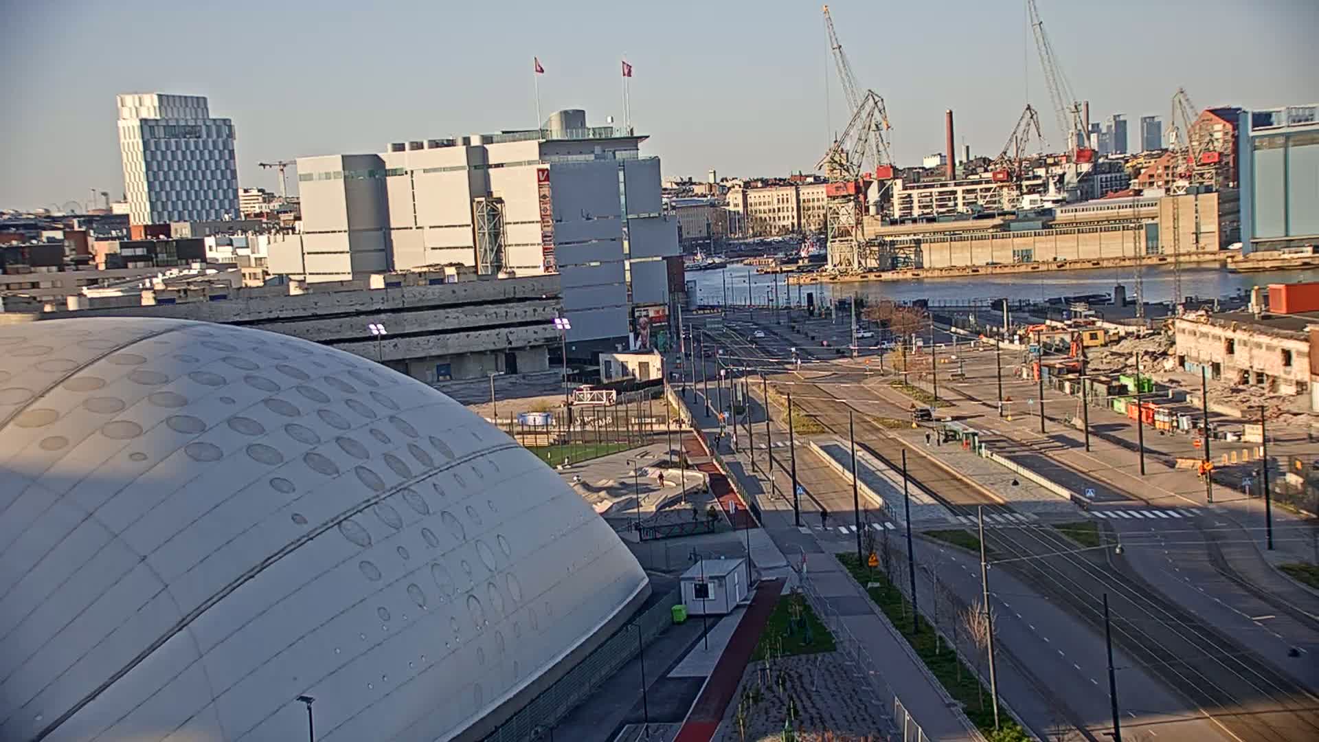 A large domed building sits adjacent to a city street with tram lines, overlooking a waterfront area with cranes and industrial buildings under a clear, sunny sky.