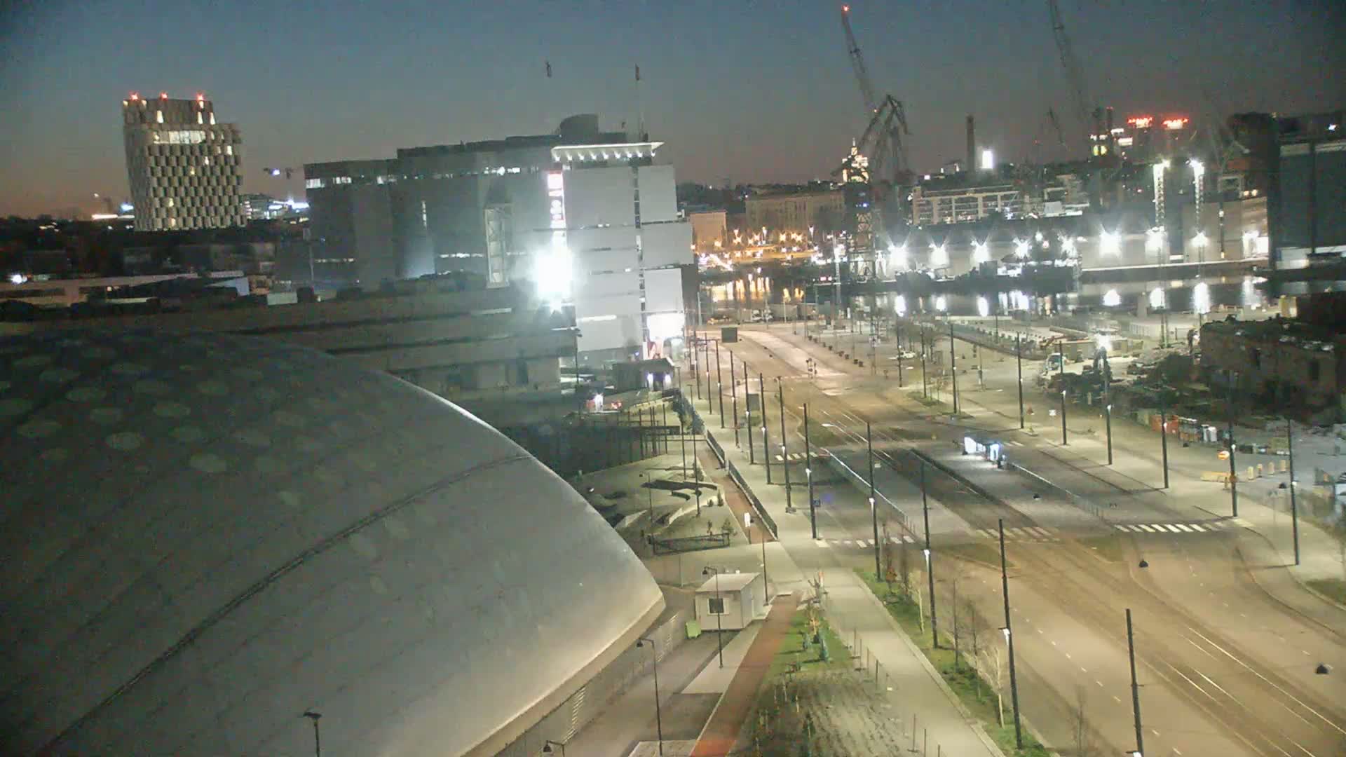 A nighttime, high-angle view shows a city street scene with a large domed building in the foreground, several other buildings in the background, and construction cranes visible in the distance.