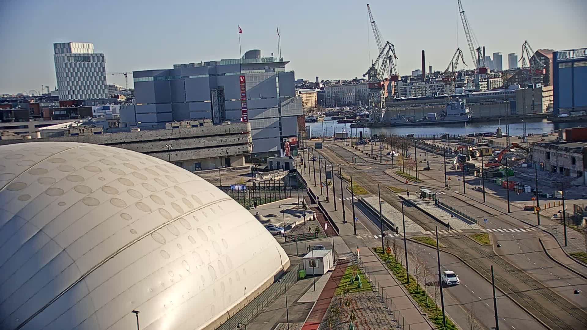 A large, domed building sits beside a wide, mostly empty road leading to a harbor filled with ships and cranes under a clear, sunny sky.