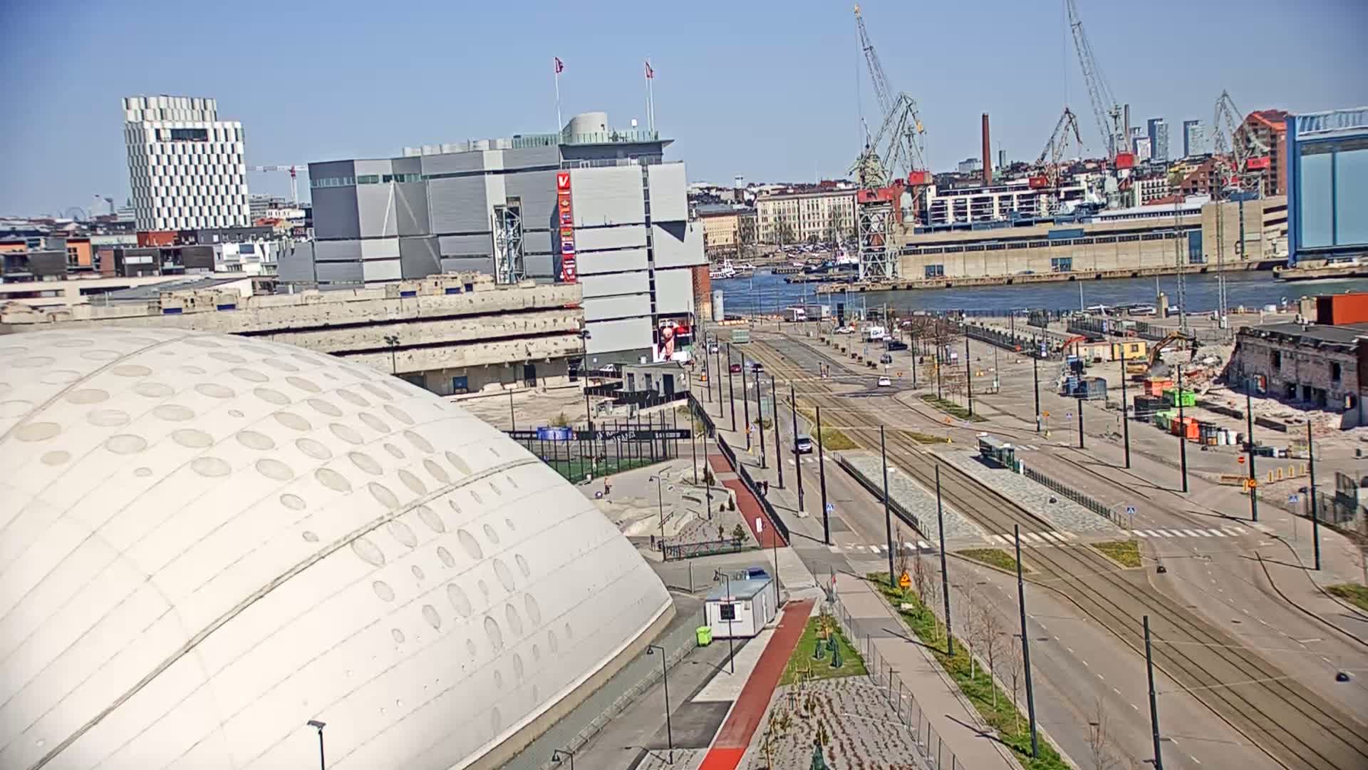 A large domed building is situated near a waterfront area with a roadway, tram tracks, and several buildings, under sunny skies.