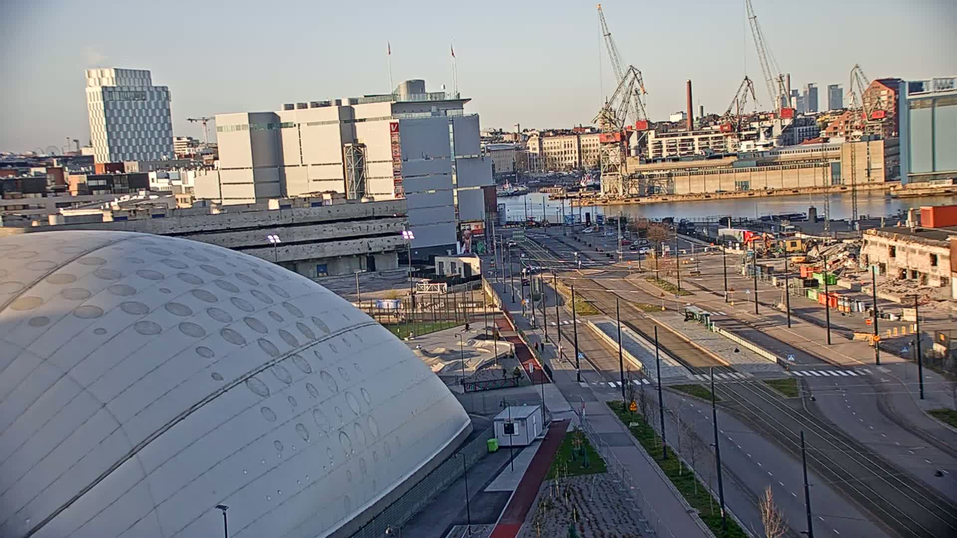 A high-angle view of a city harbor on a sunny day shows a large domed building, streets with tram tracks, industrial areas with cranes, and buildings under demolition.