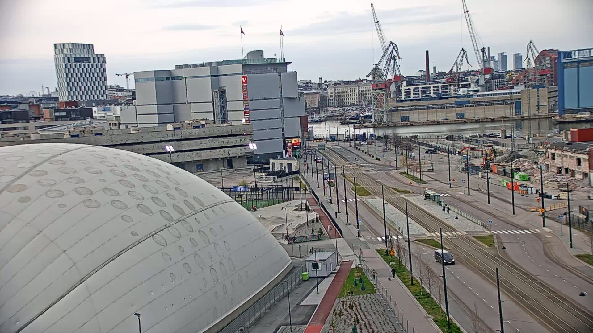 A large domed building sits beside a wide street with tram tracks running through it, under a cloudy sky, with a waterfront and several cranes in the background.