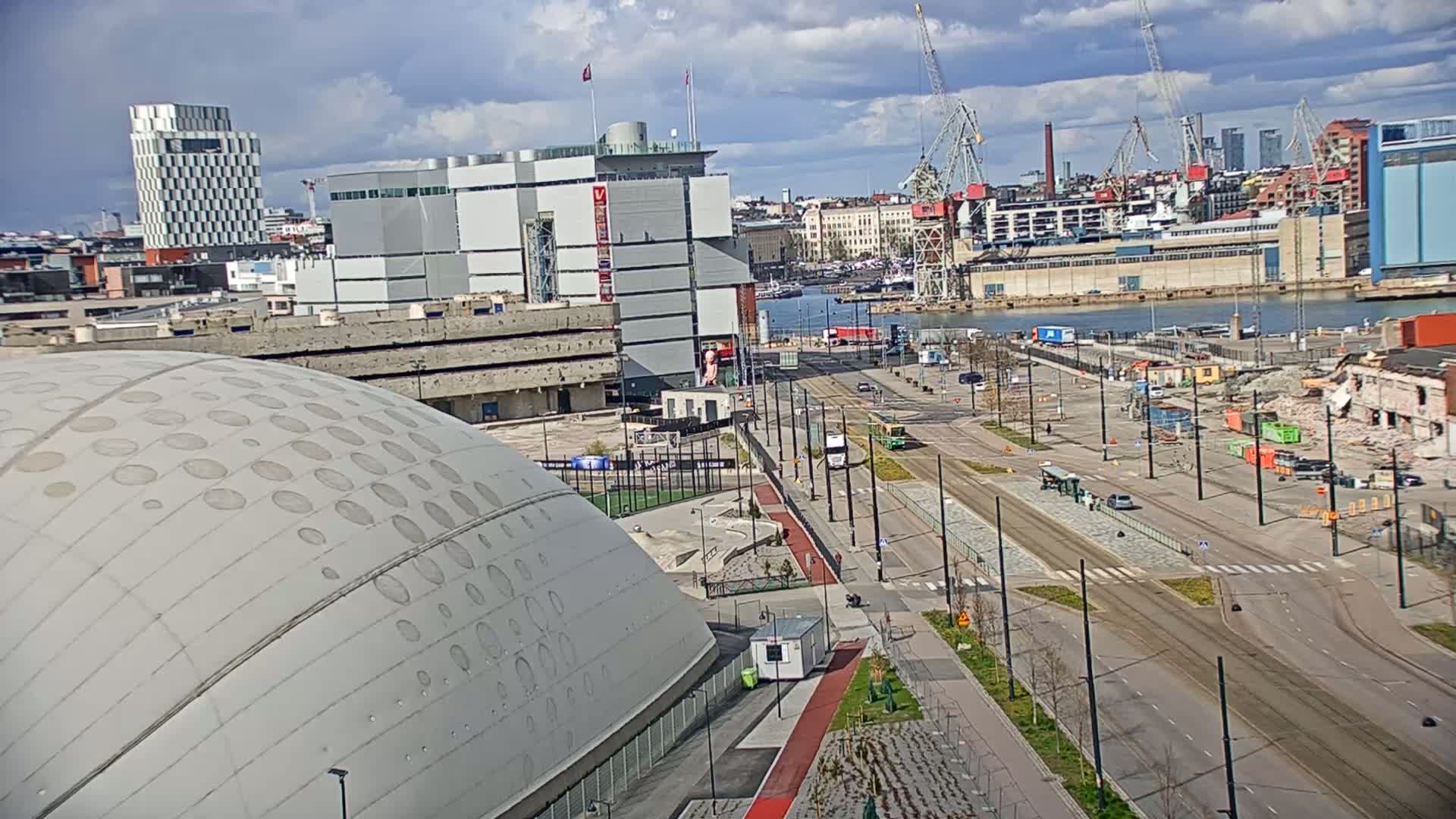 A partly cloudy day reveals a cityscape with a large domed building in the foreground, a waterfront with active construction and cranes, and several other buildings stretching into the distance.