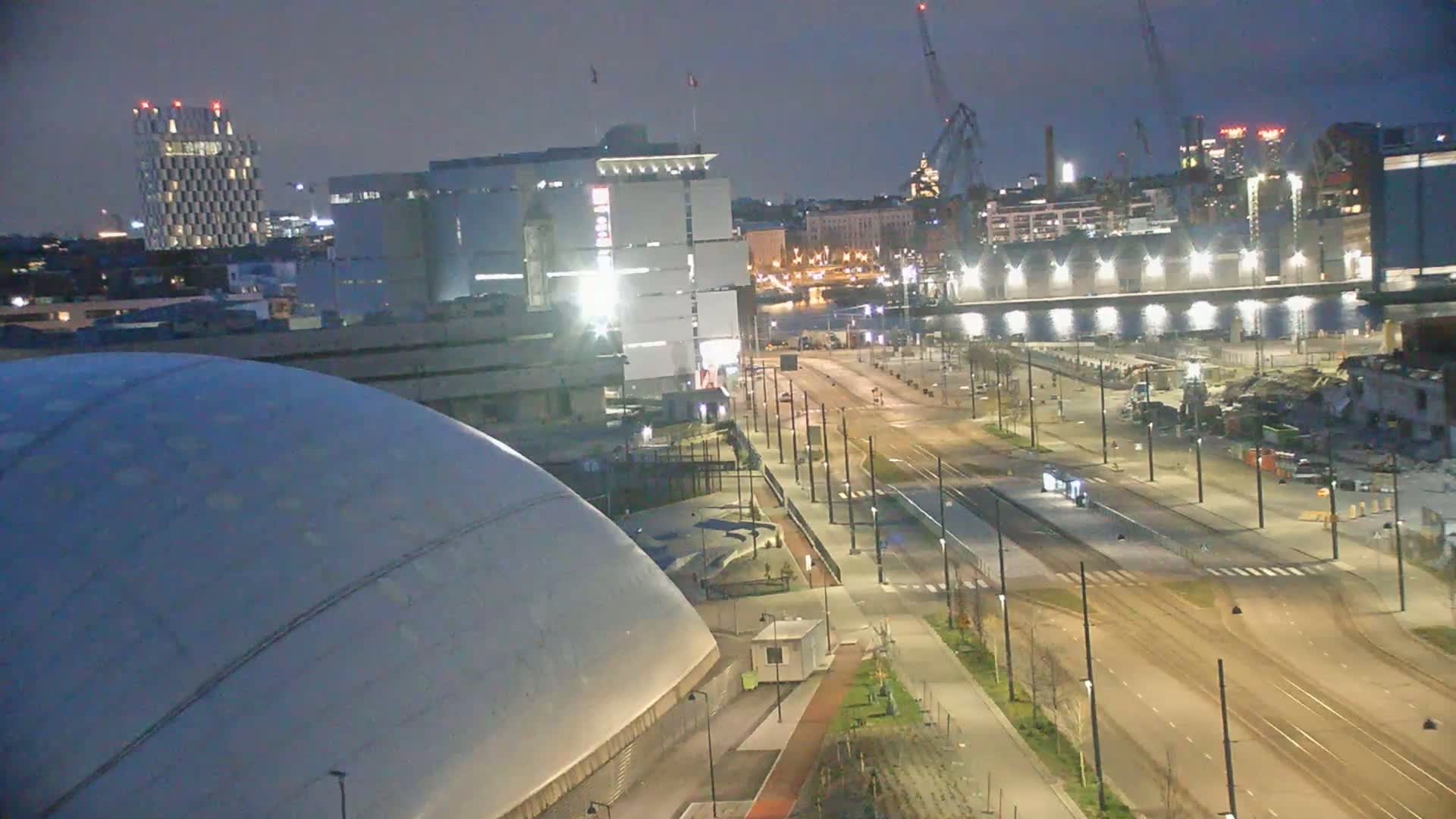 A nighttime view of a city featuring a large, domed building, several other buildings, and a long, empty street with tram tracks, under a dark sky.