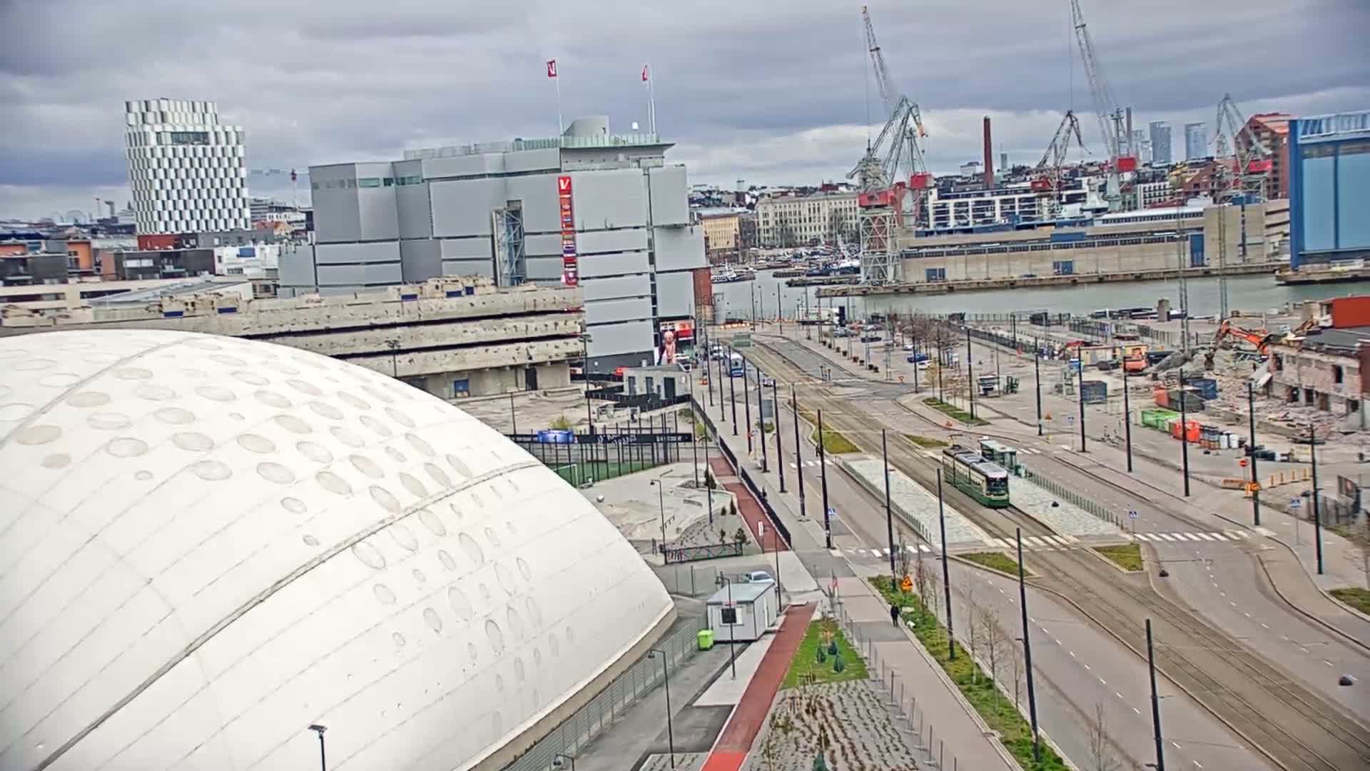 A large white domed building sits beside a city street with a tram, under a cloudy sky, overlooking a harbor with construction cranes and demolition activity.