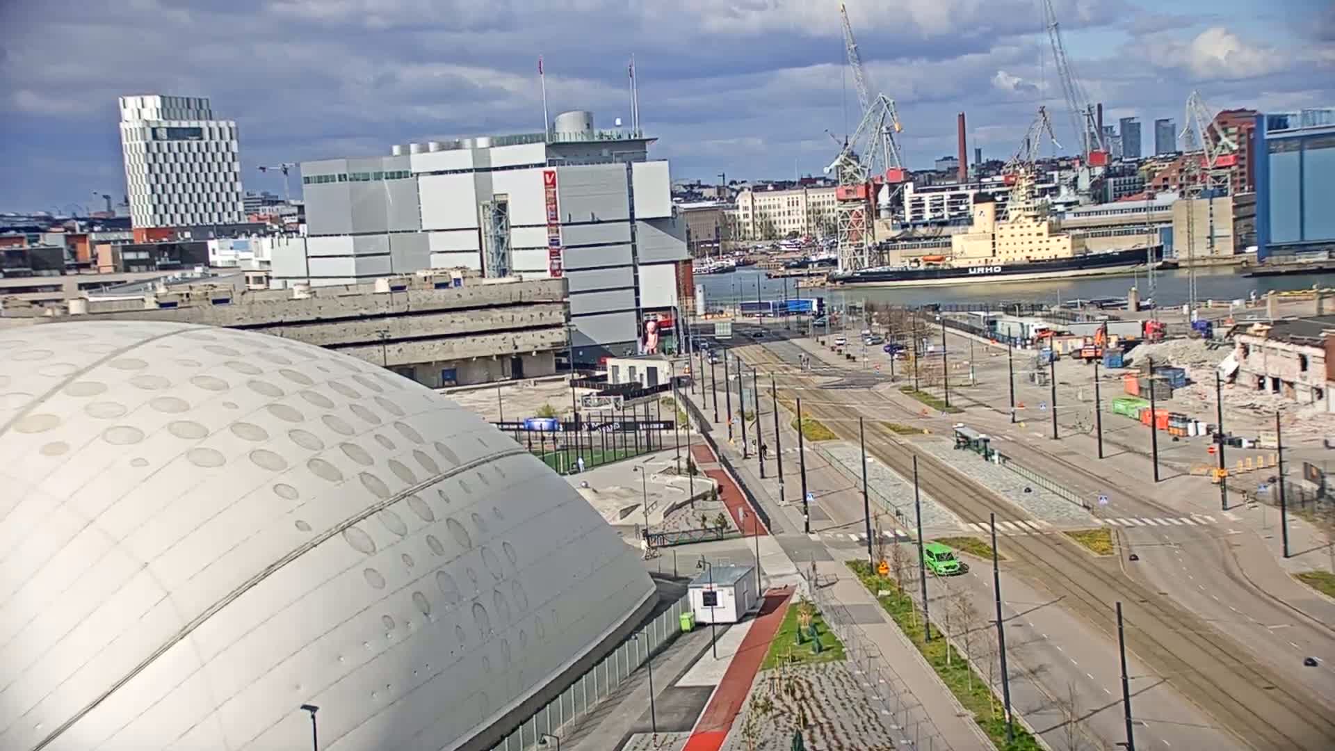 A partly cloudy day reveals a cityscape with a large domed building in the foreground, a harbor with a ship, and ongoing construction or demolition in the midground.