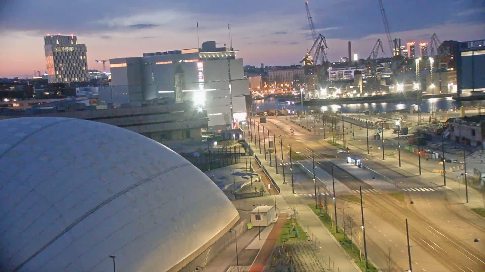 An urban cityscape at twilight shows a large domed building in the foreground, a wide street with tram lines, and a harbor with cranes in the background under a mostly clear sky.